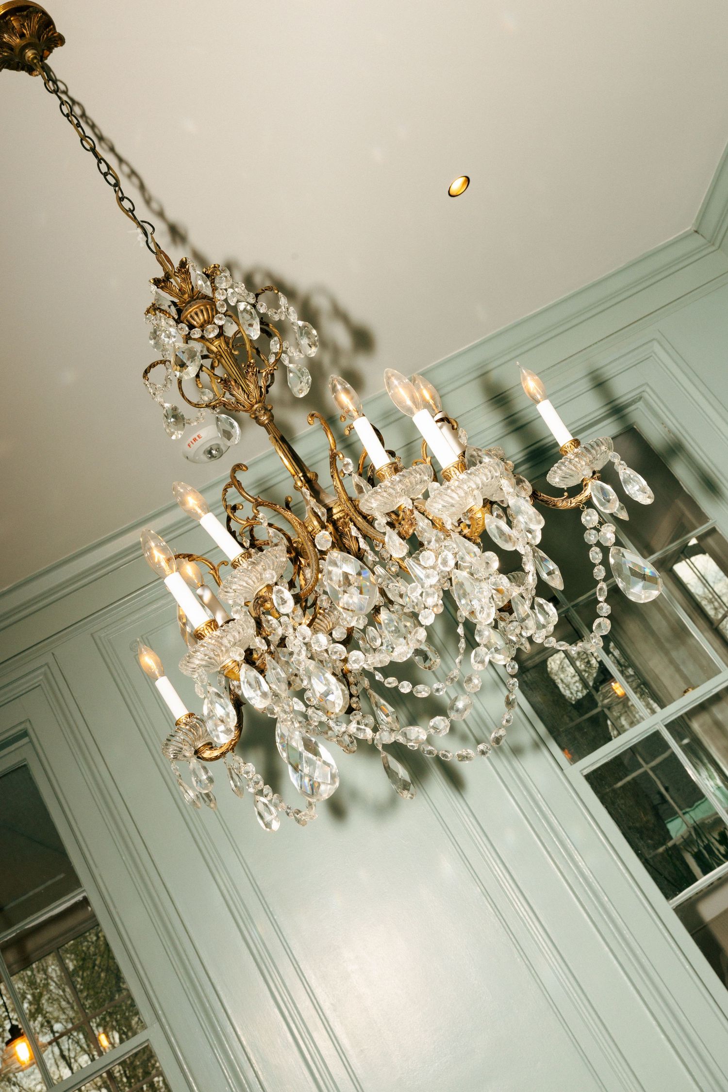 Elegant crystal chandelier with ornate gold metalwork hangs from a mint green ceiling in a classic interior space.