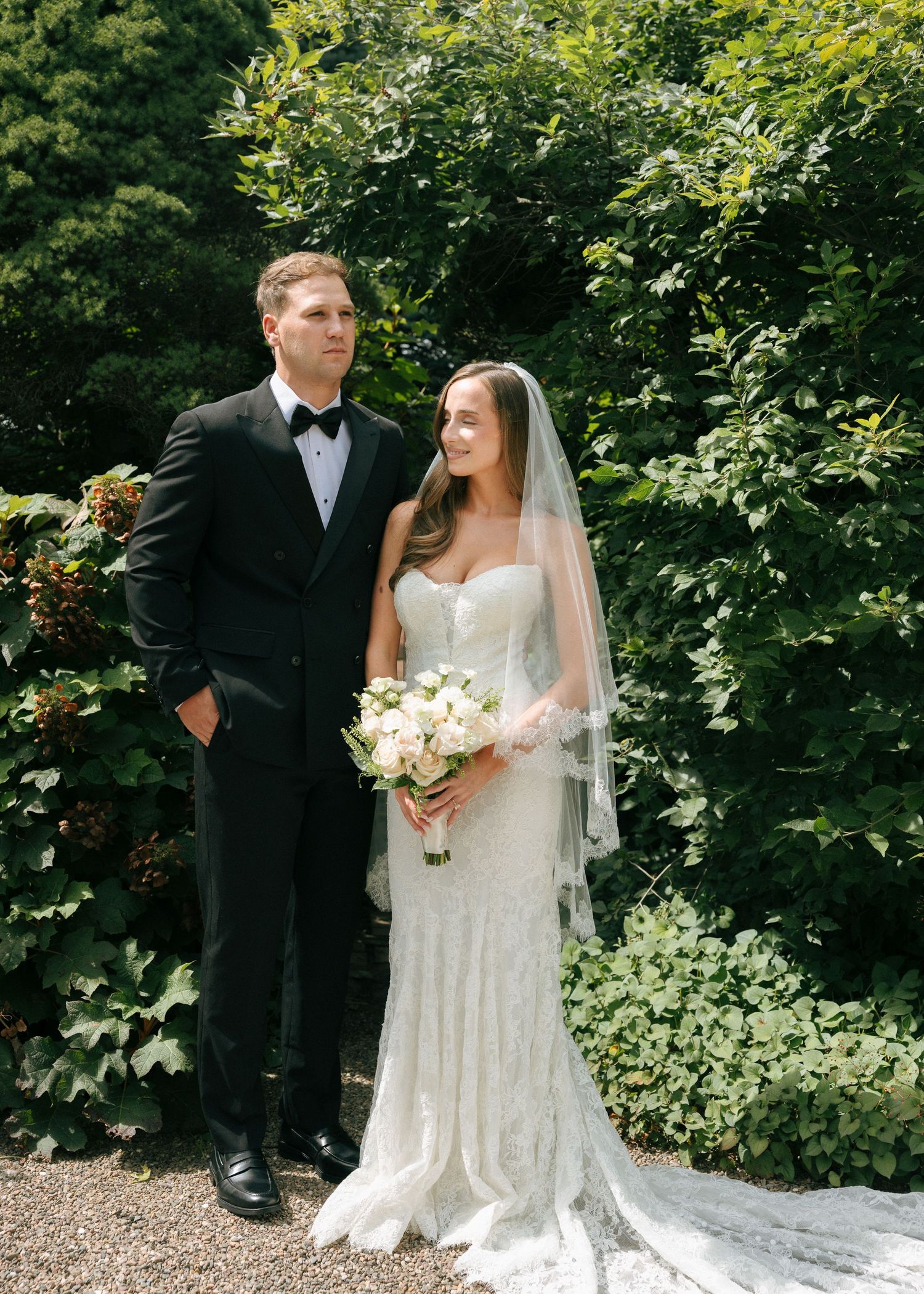 Bride and groom pose together in a lush garden, she holds white bouquet in beaded gown, he wears classic black tuxedo.