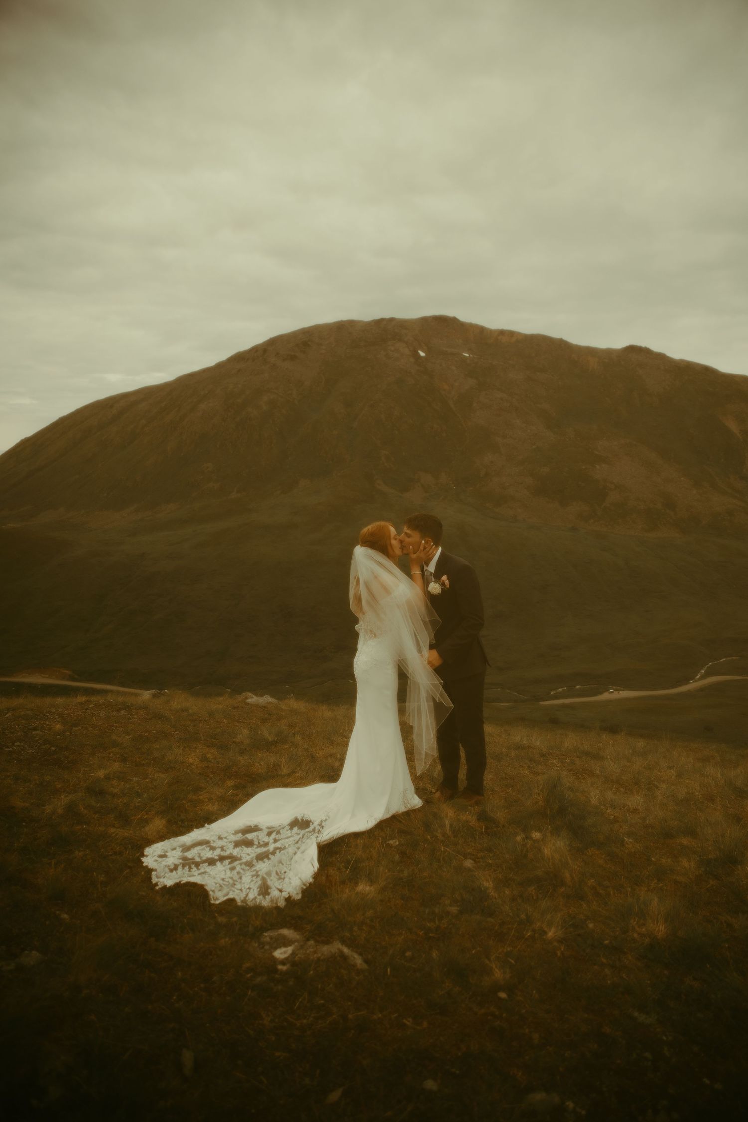 A couple embraces on a misty hillside in moody lighting with a dramatic mountain backdrop and long white wedding dress train.
