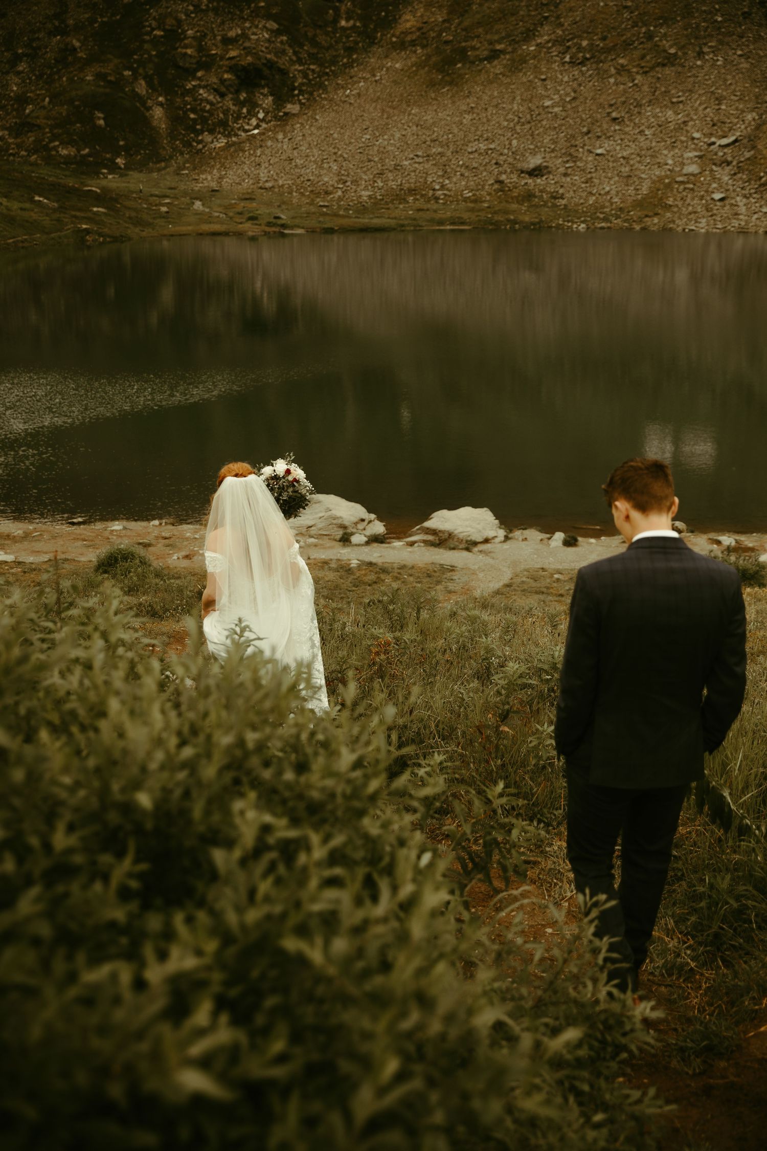 A couple stands near a serene mountain lake surrounded by natural terrain on their wedding day.