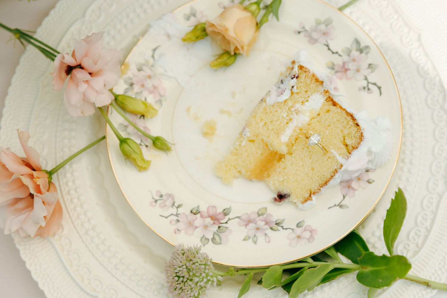 A slice of vanilla cake sits on a floral-patterned plate surrounded by pink flowers and greenery.