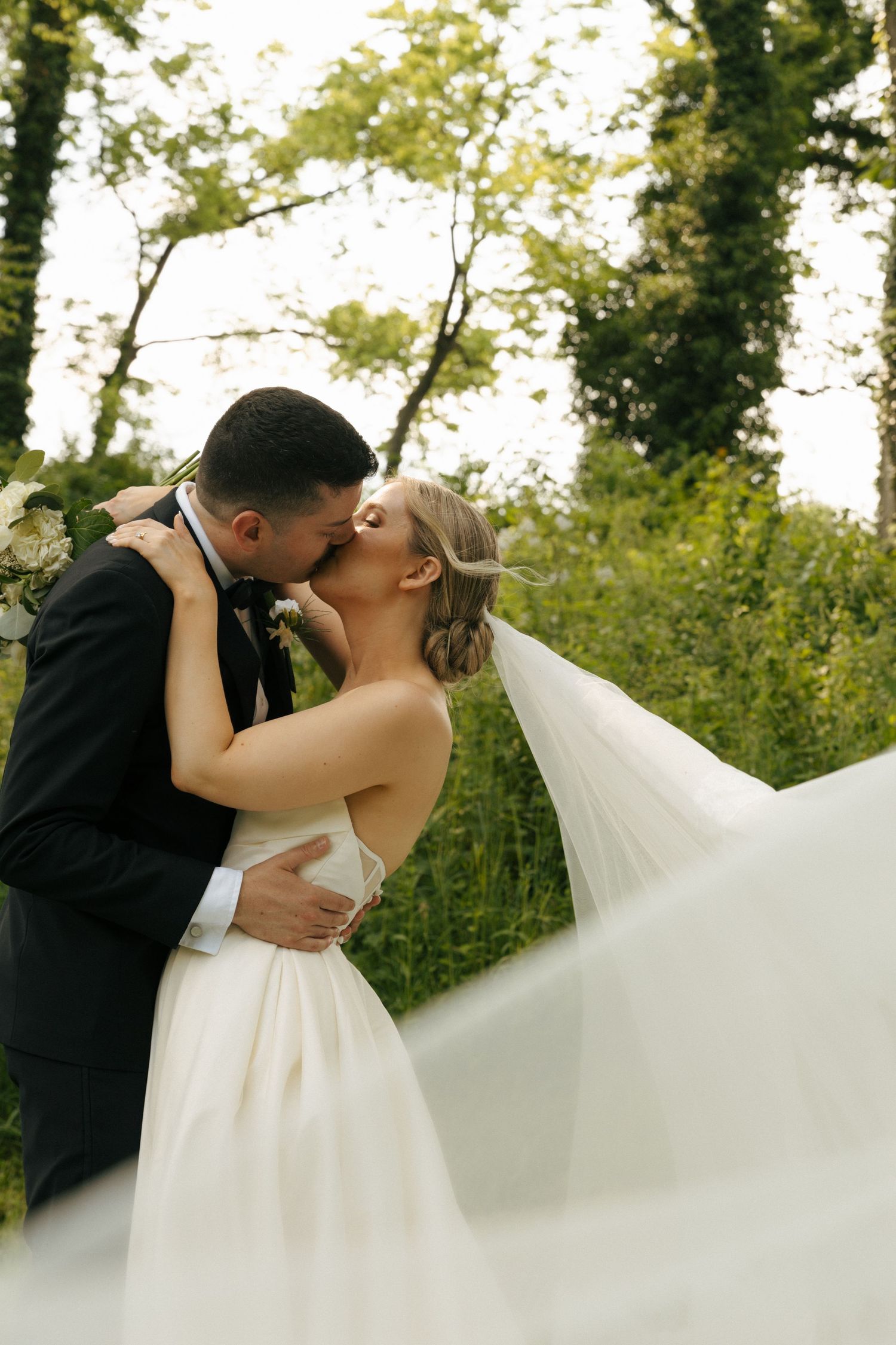 A couple sharing an intimate kiss in their wedding attire with a flowing veil catching the breeze in a natural outdoor setting.