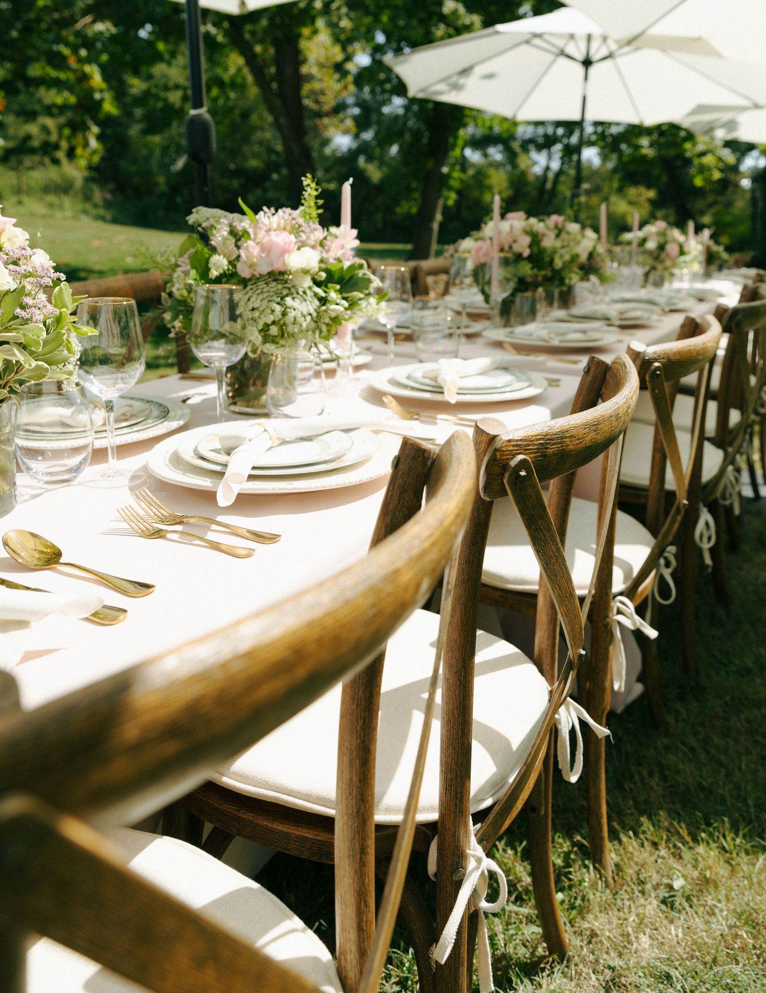 Elegant outdoor table setting with rustic wooden chairs, white linens, and soft pink floral centerpieces under garden umbrellas.
