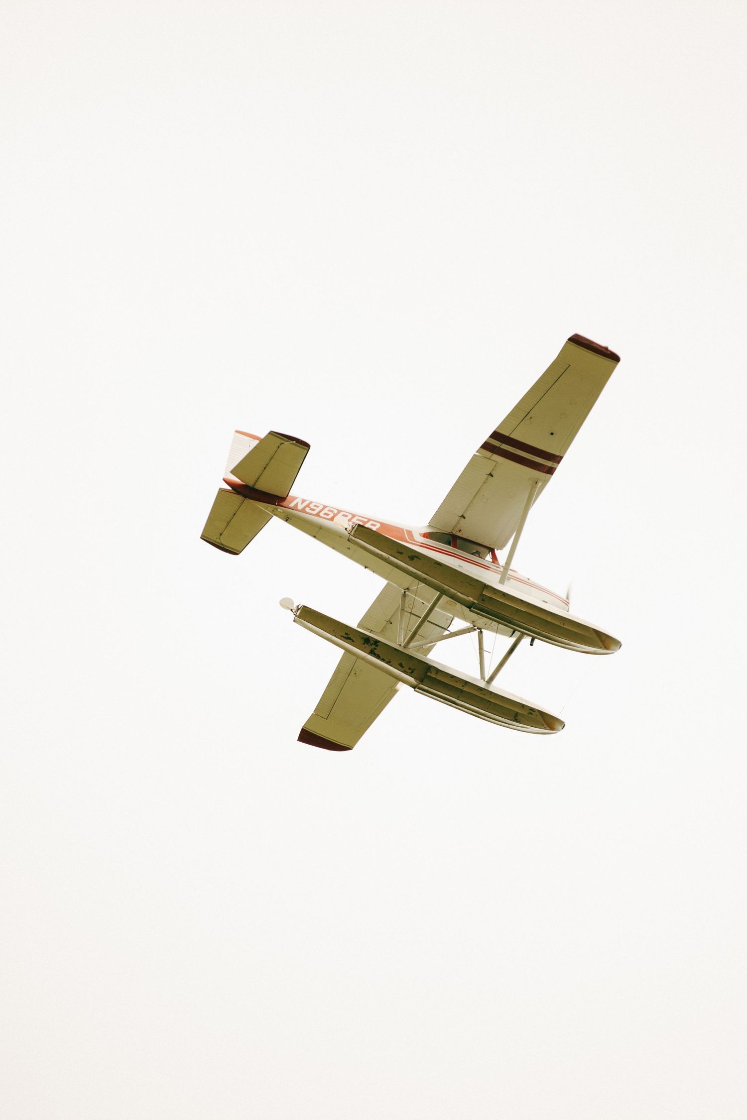 A seaplane with floats photographed from below against a white sky mid-flight.