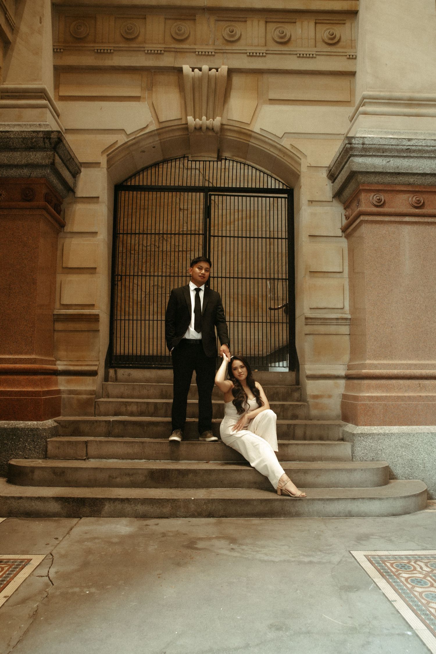 Couple in formal attire posed on vintage stone steps by ornate iron gate and architectural details of historic building.