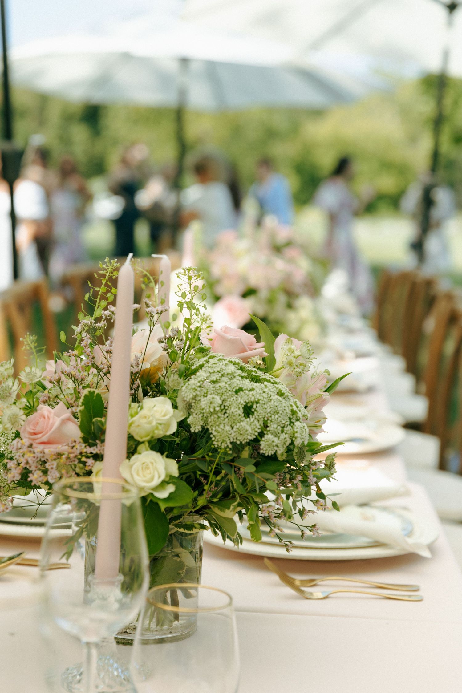 Elegant outdoor wedding table setting with white and pink floral centerpieces and delicate place settings under a tent.