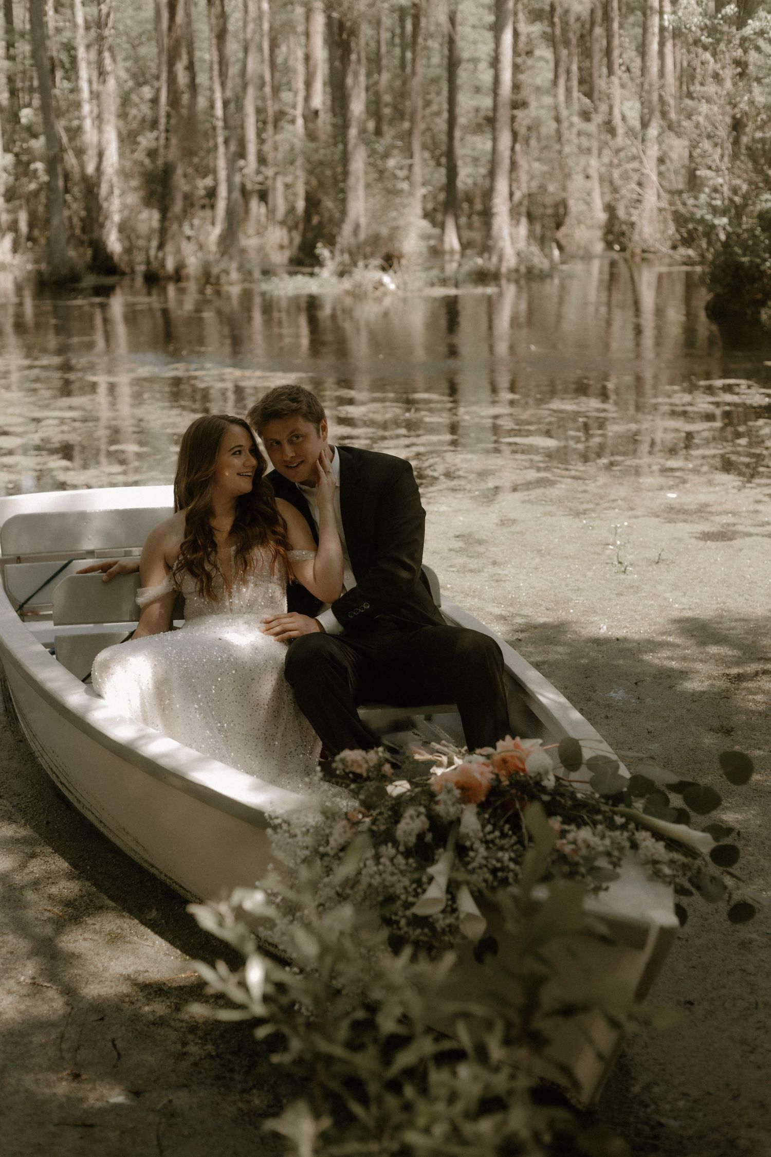 Romantic wedding couple sits in vintage rowboat surrounded by cypress trees and reflective water in ethereal swamp setting.
