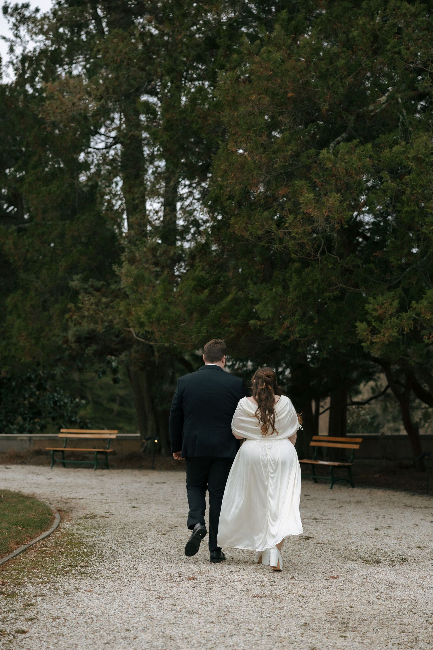 A couple in wedding attire walks away on a gravel path surrounded by trees in an outdoor setting.