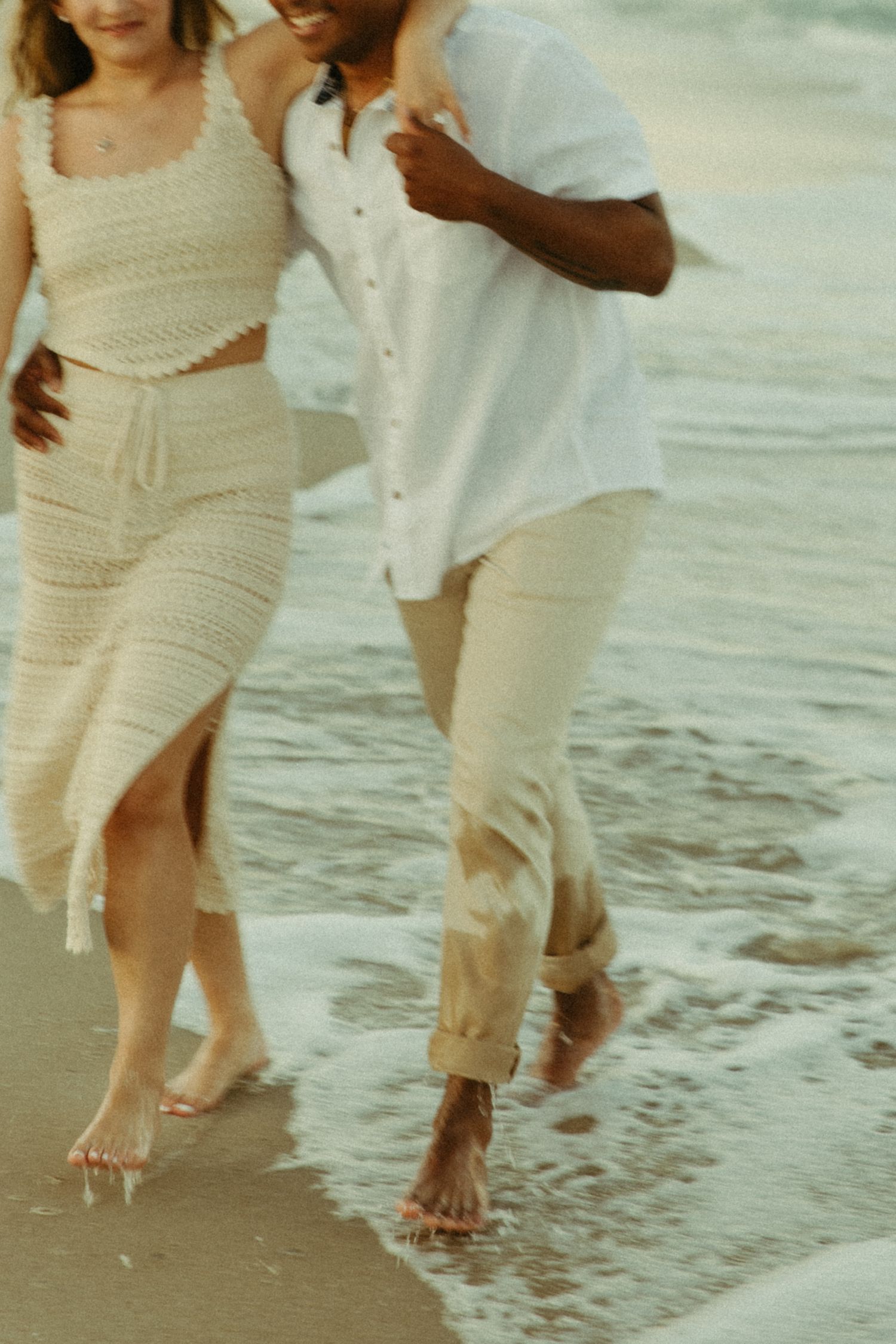 A romantic couple walking barefoot along the beach shoreline wearing white and cream colored summer outfits.