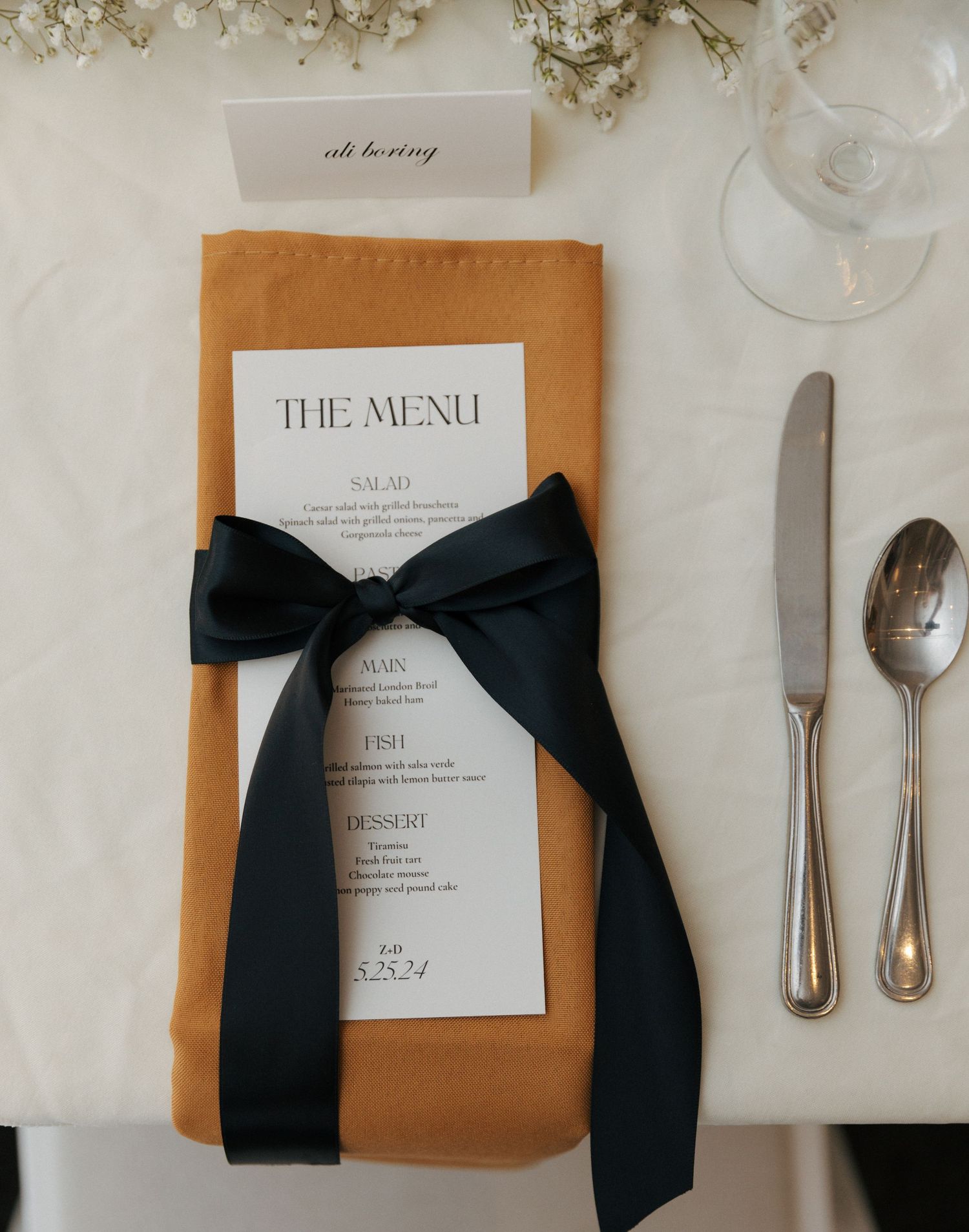 Elegant place setting with menu wrapped in tan napkin and black ribbon bow beside silverware on white tablecloth.