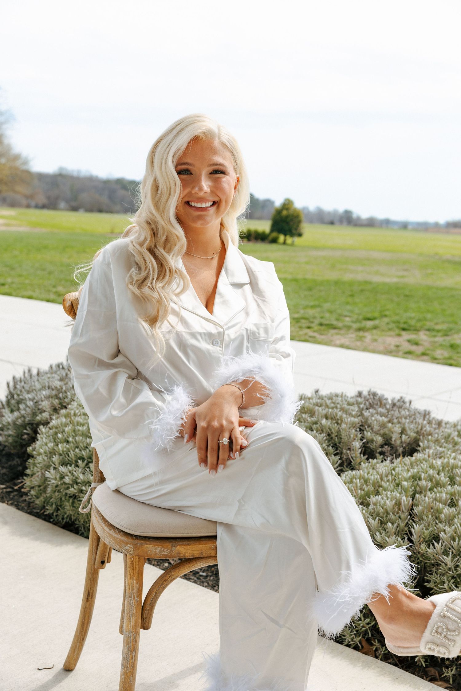 Person in white outfit sits on wooden stool outdoors near lavender plants on concrete patio with green field in background.