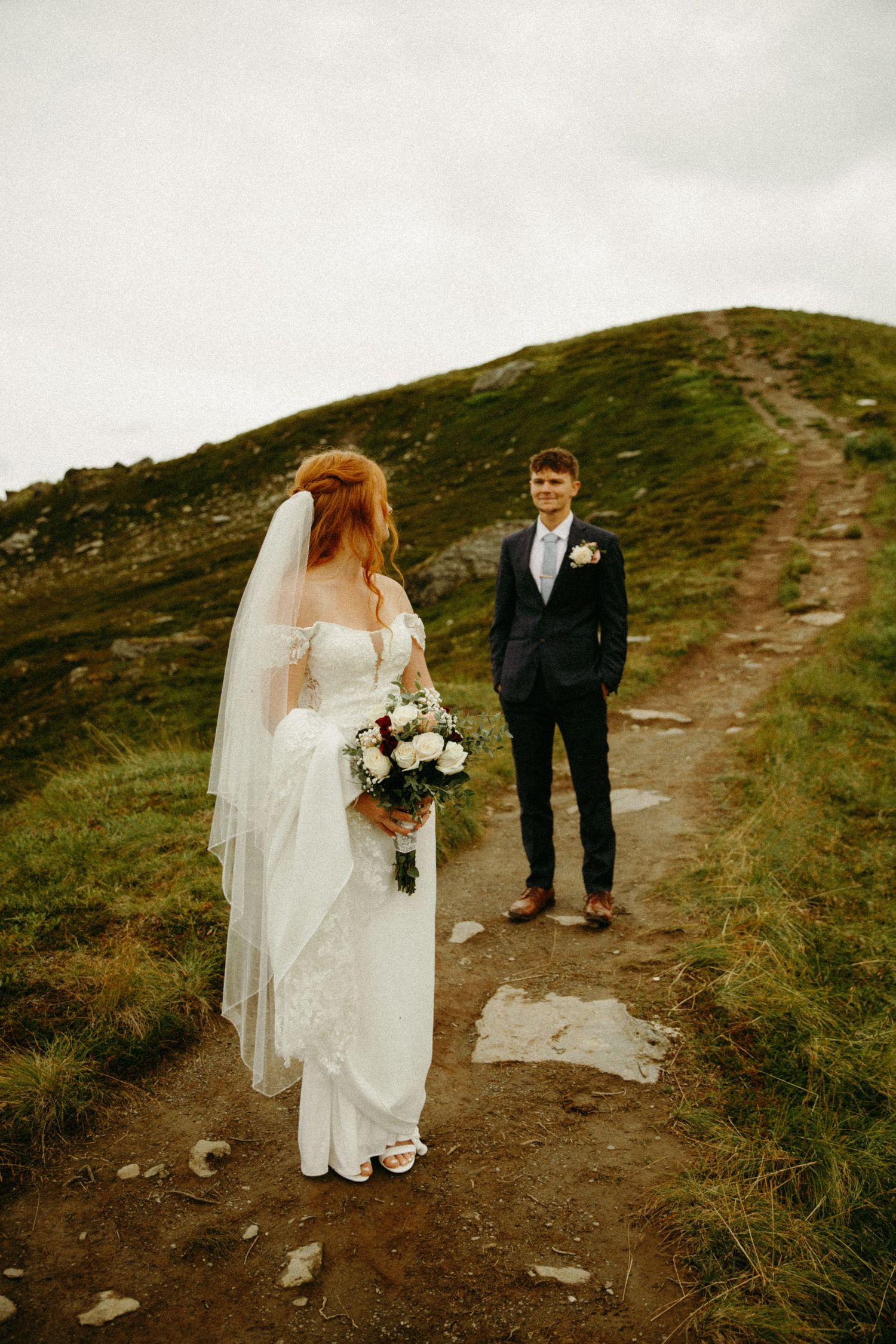 A couple stands on a scenic mountain trail during their wedding day, with green hillsides and moody skies in the background.