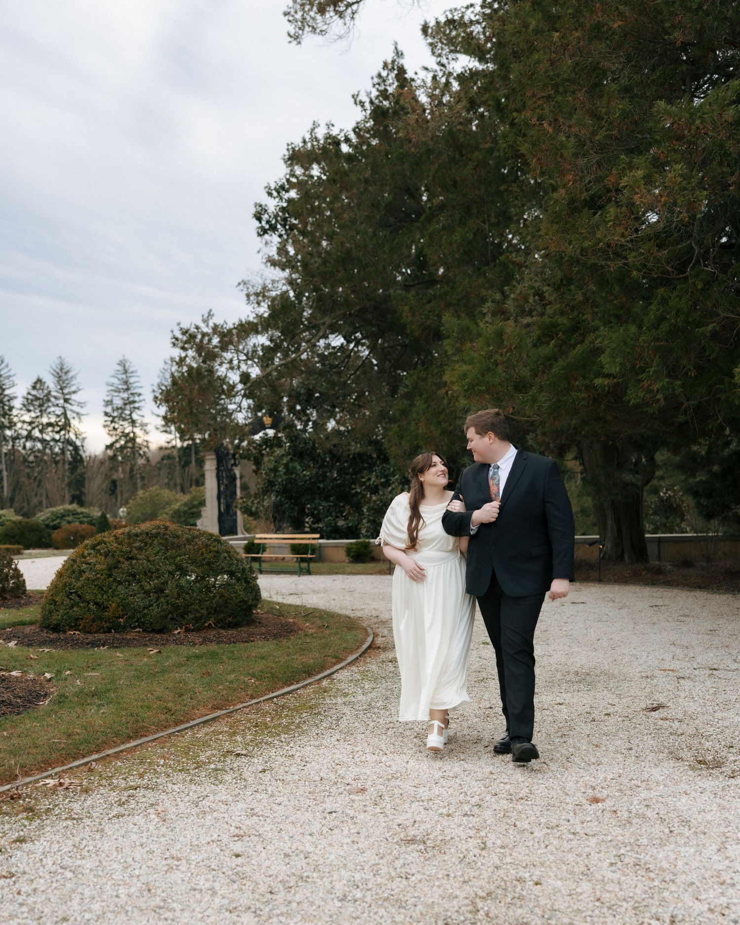 A couple in wedding attire walks along a winding gravel path surrounded by lush trees and greenery.