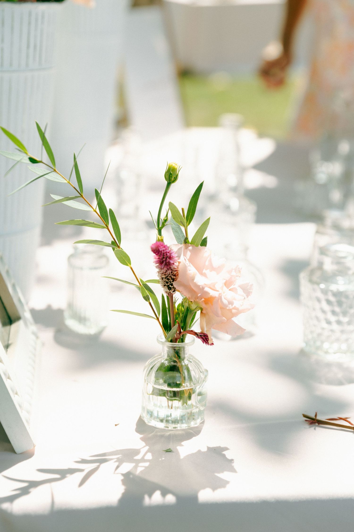 A delicate flower arrangement with pink blooms and greenery in a small glass vase on a sunlit white table.