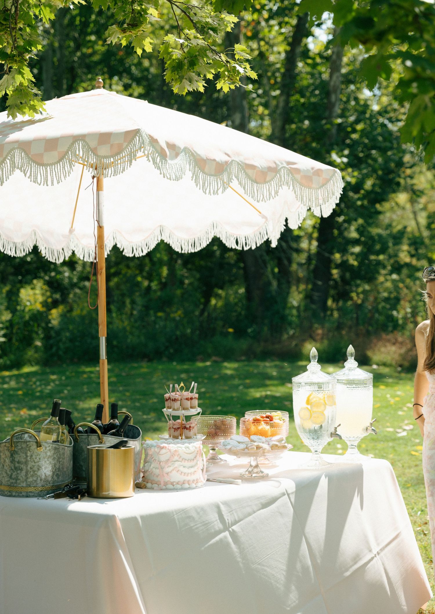 An elegant garden party setup with a vintage fringed umbrella over a white table with refreshments and glass drink dispensers.