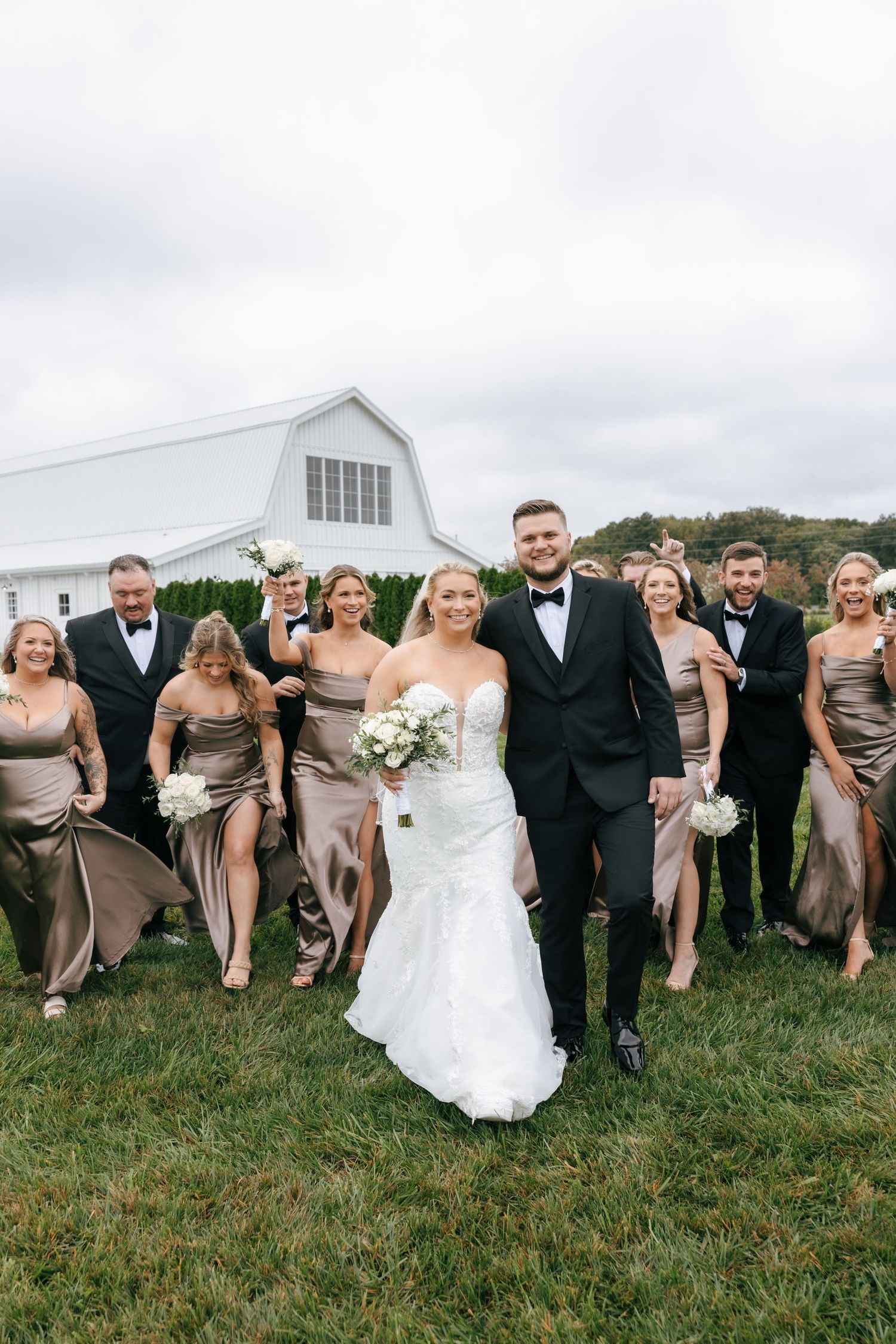 Wedding party in formal attire poses playfully on grassy hillside with white barn in background.