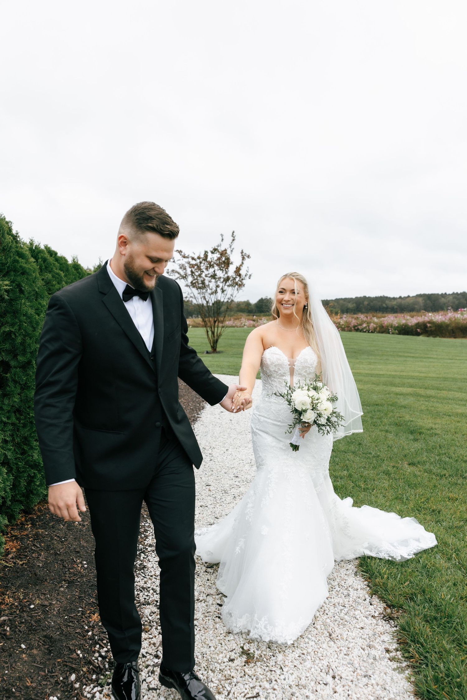 A bride and groom share candid moments while walking along a gravel path surrounded by green fields on their wedding day.