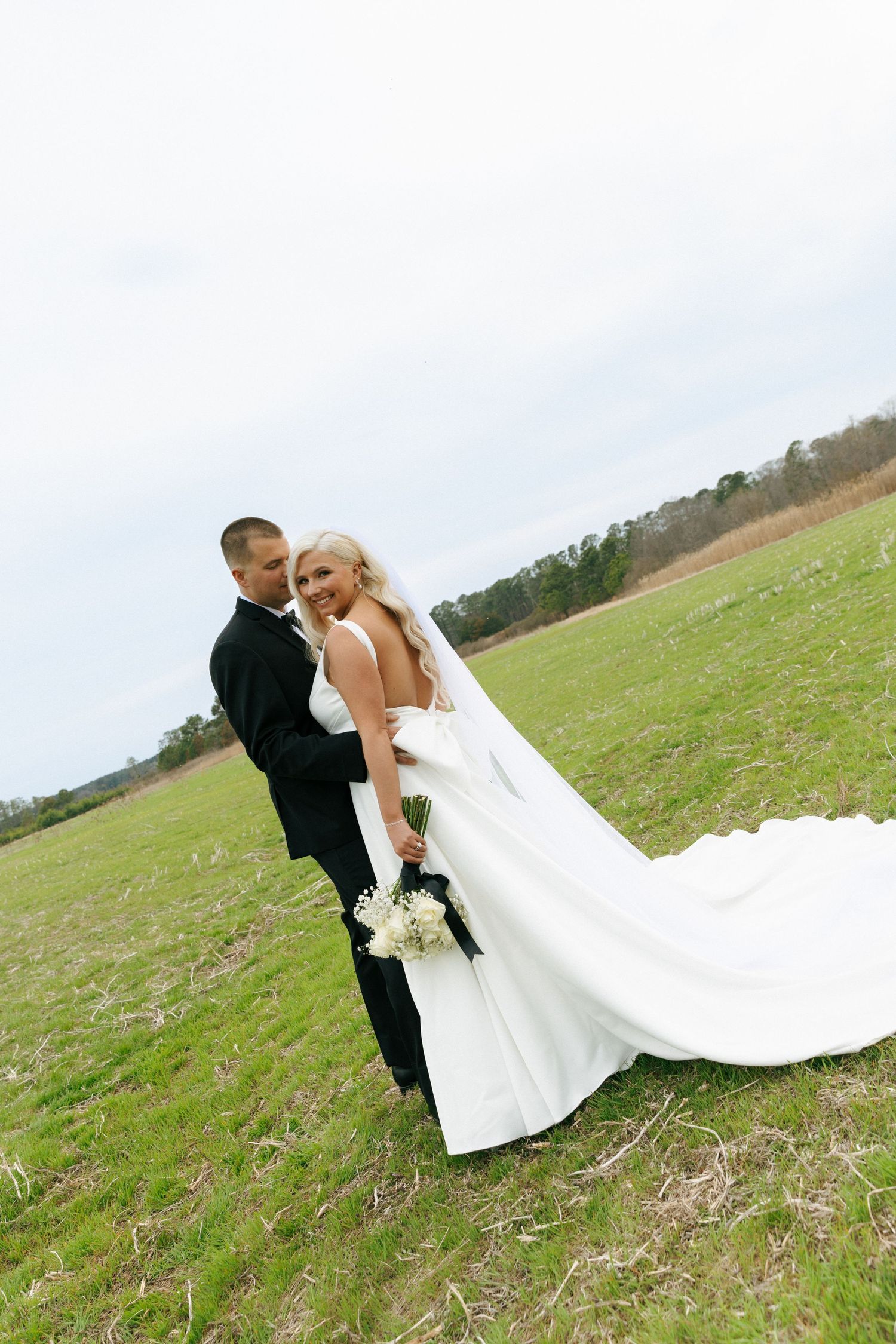 A couple in wedding attire embrace on a grassy hillside with a long white veil flowing in the breeze.