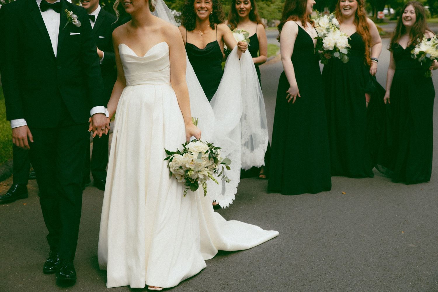 Wedding party with bride in white gown and bridesmaids in black dresses standing together on asphalt.
