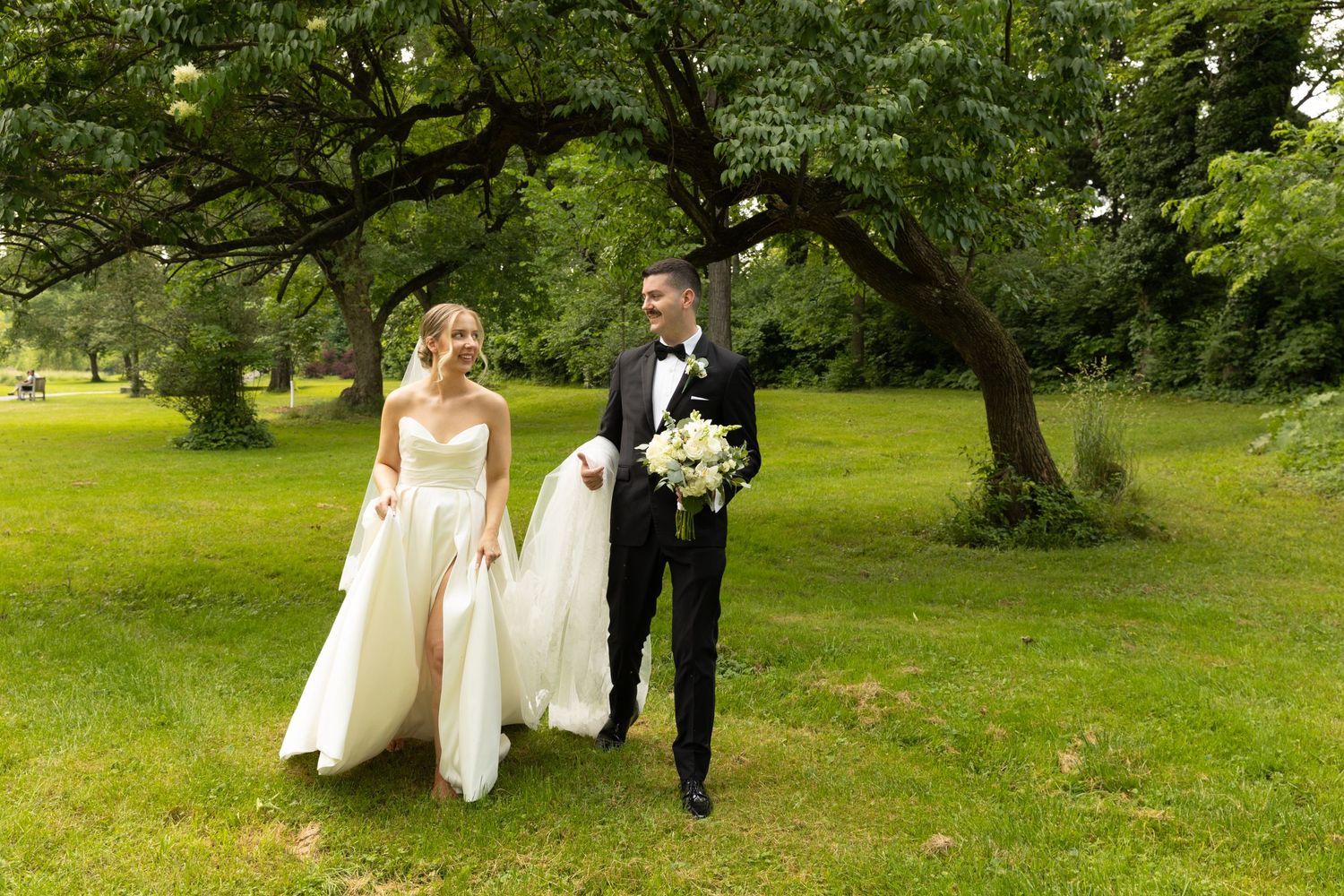 A romantic wedding couple walks together on a lush green lawn under mature trees on a sunny summer day.