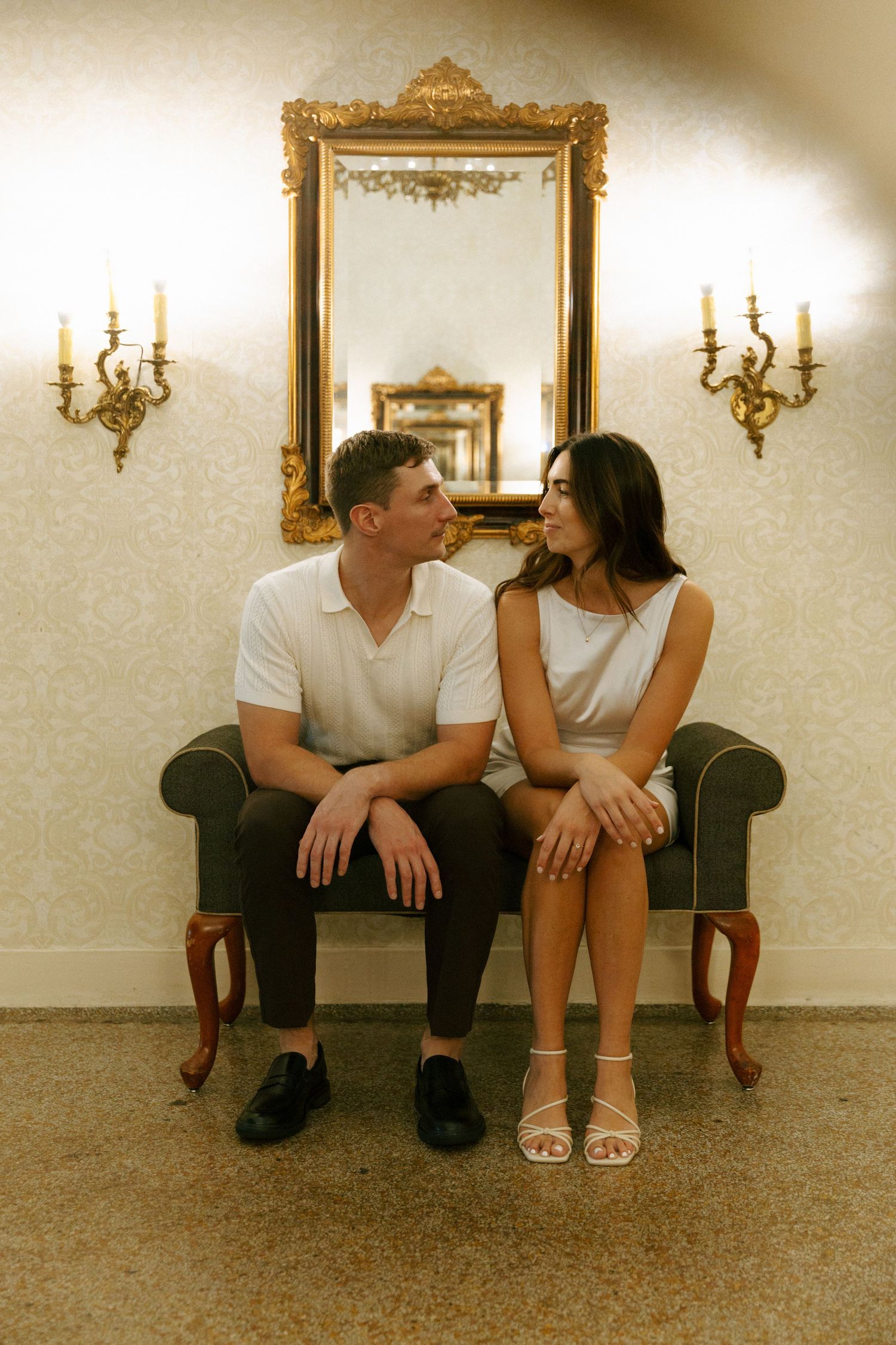 Couple sitting together on vintage loveseat with ornate gold mirror and wall sconces in elegant interior.