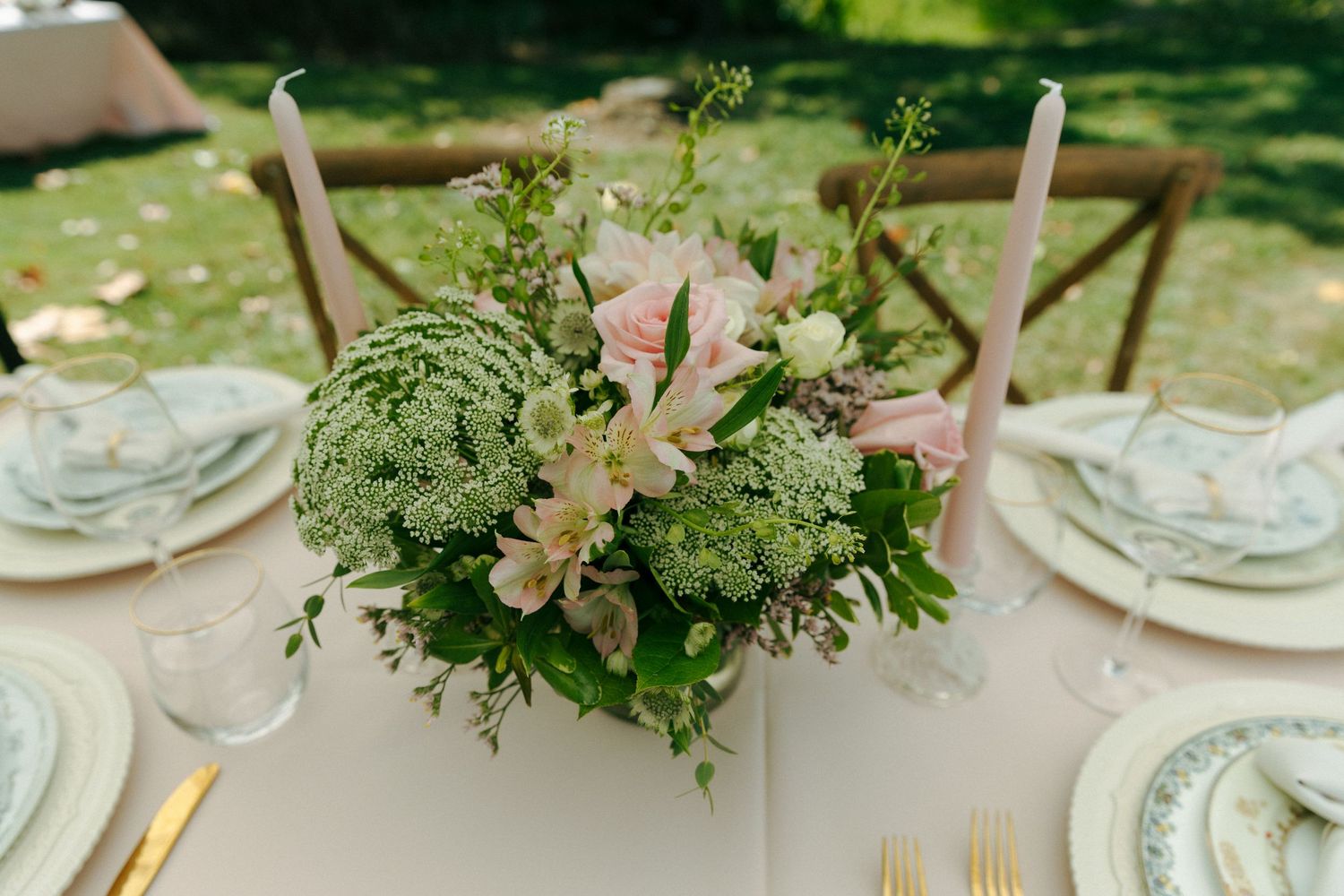 An elegant outdoor wedding reception table setting with pastel pink and green floral centerpiece and white table settings.