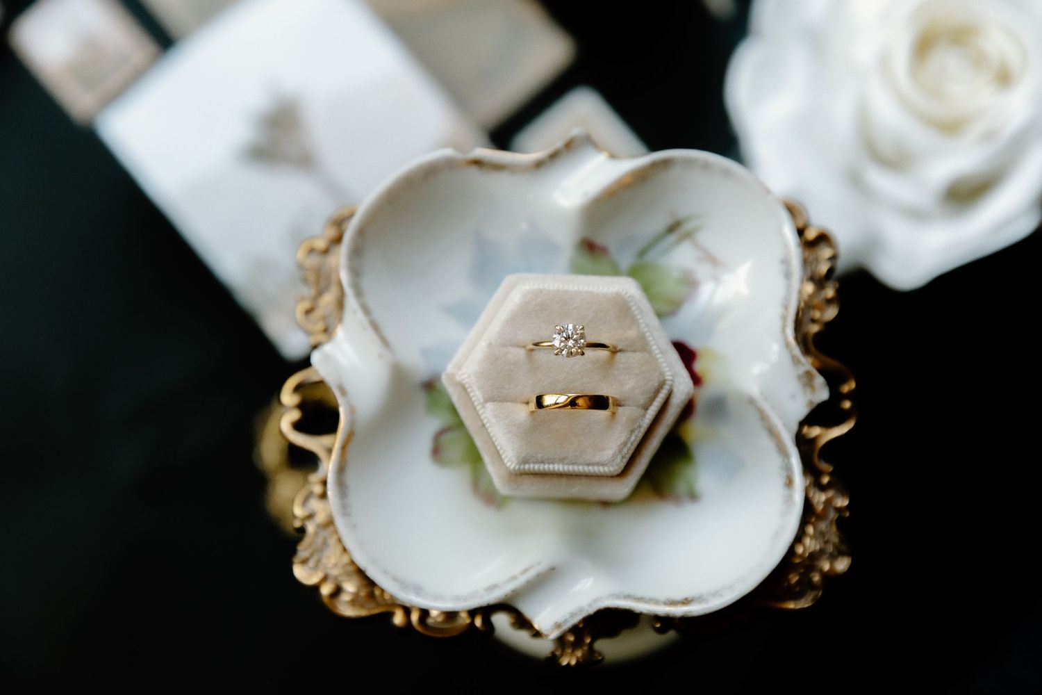 Wedding rings nestled in a white floral-shaped ring box with gold trim on dark background.