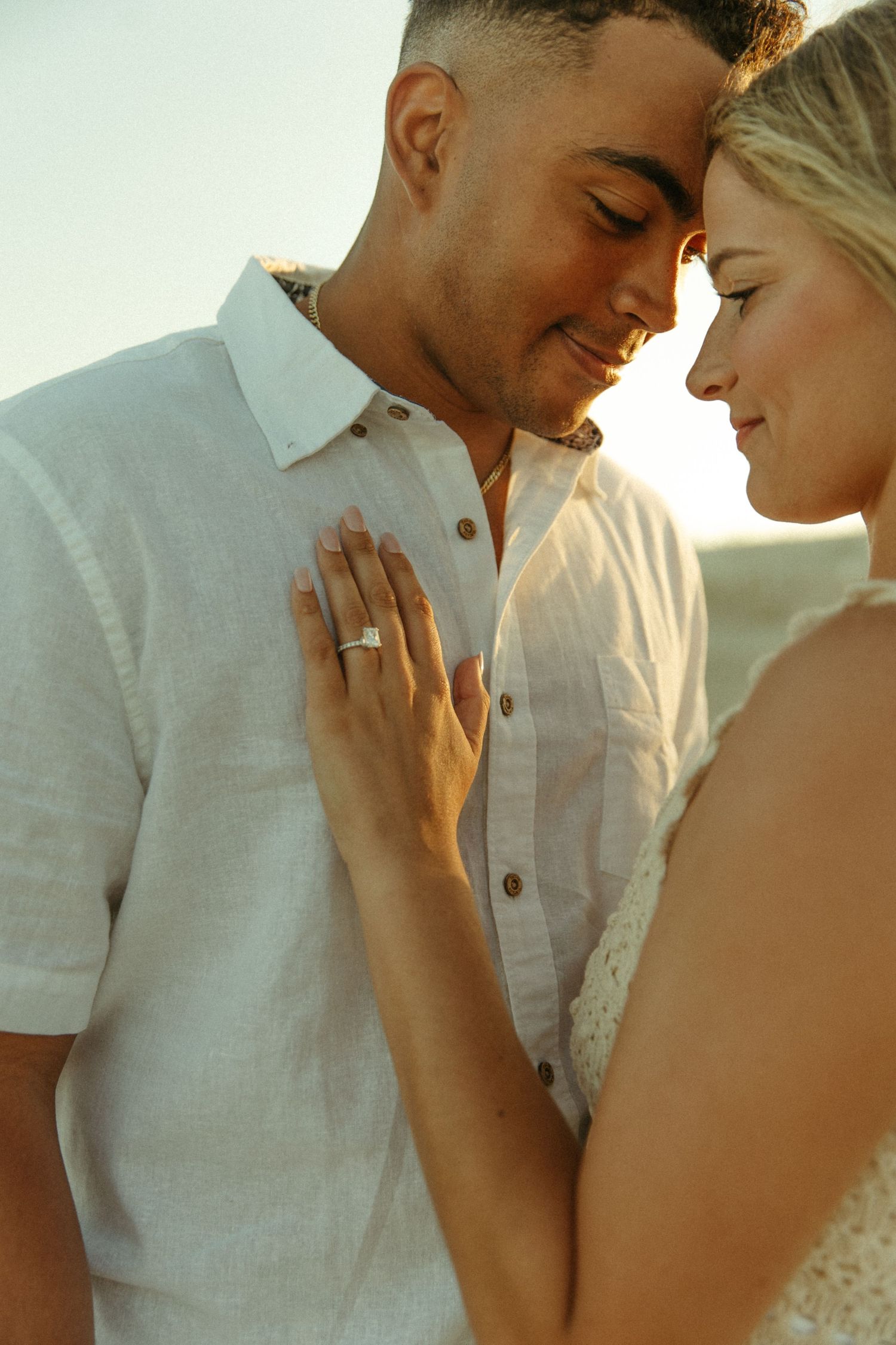 A romantic couple embraces in warm sunlight, with her hand resting on his white shirt as they share an intimate moment.