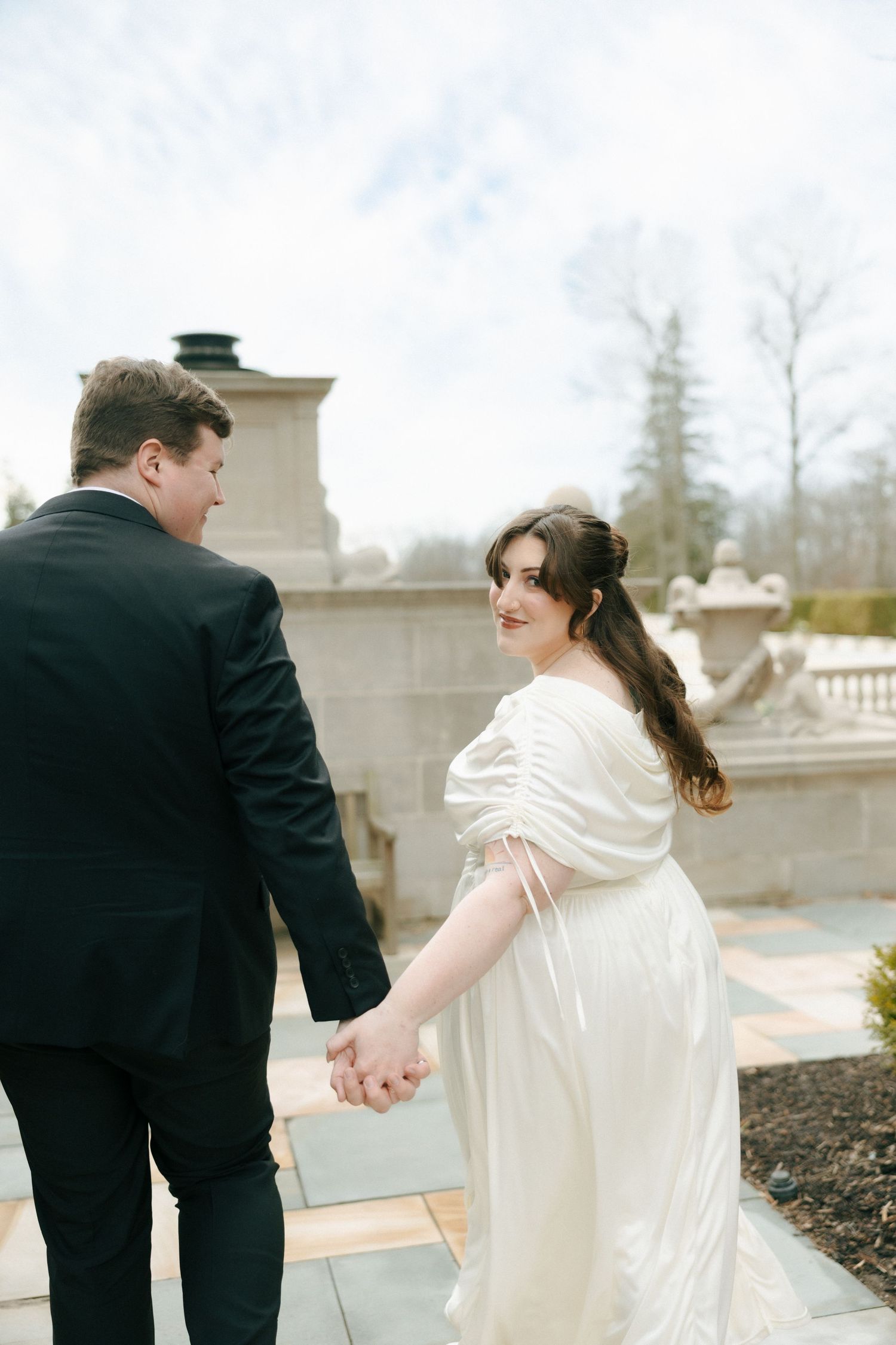 A couple in wedding attire holding hands while walking along a stone balustrade in an outdoor setting.