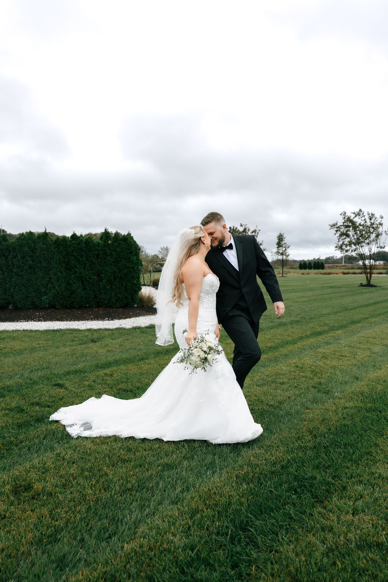 A couple in wedding attire dances together on a lush green lawn under cloudy skies.