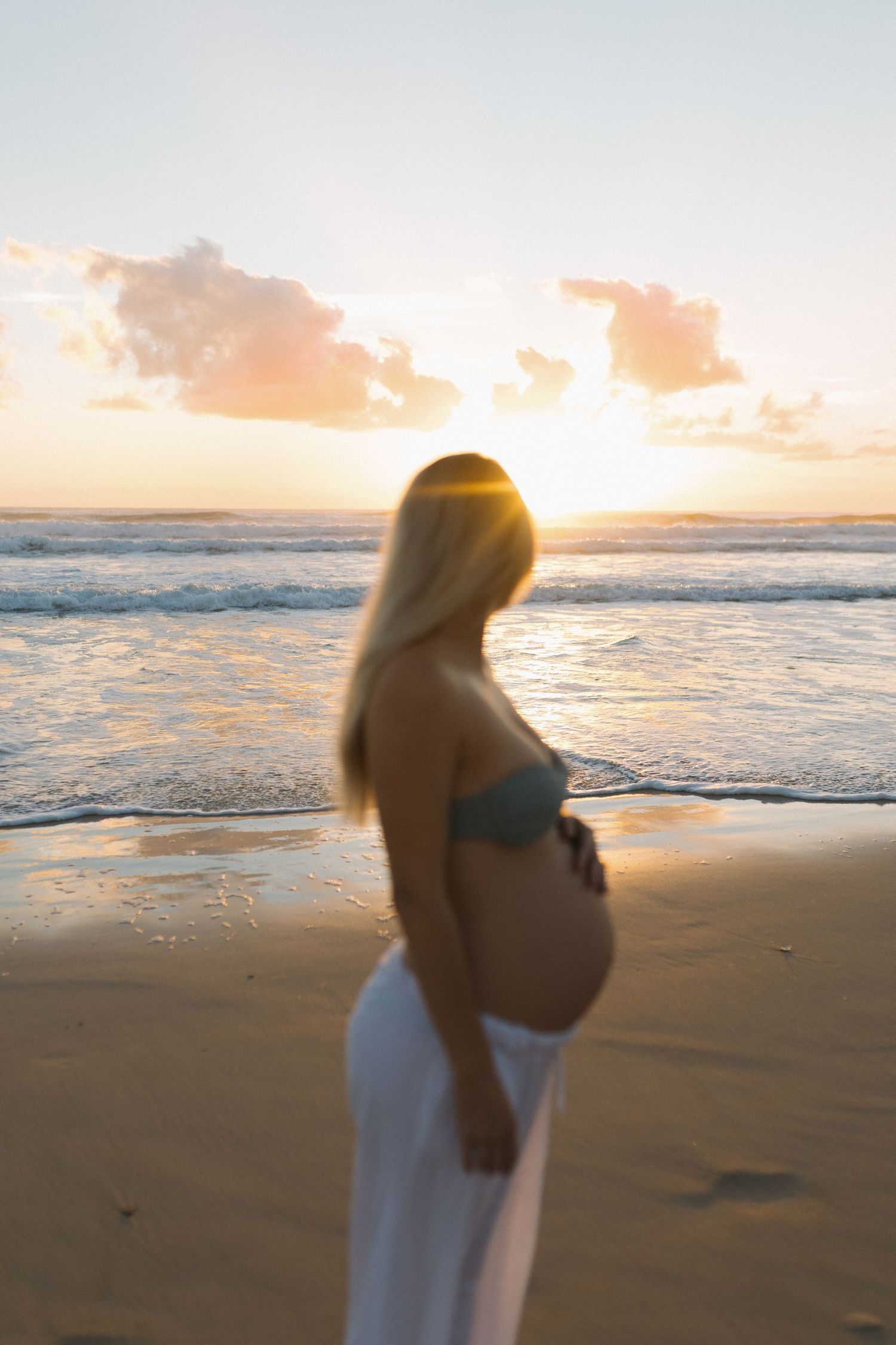 Pregnant woman in bikini top and white pants standing on beach at golden sunrise, cradling baby bump by ocean waves at the Gold Coast.