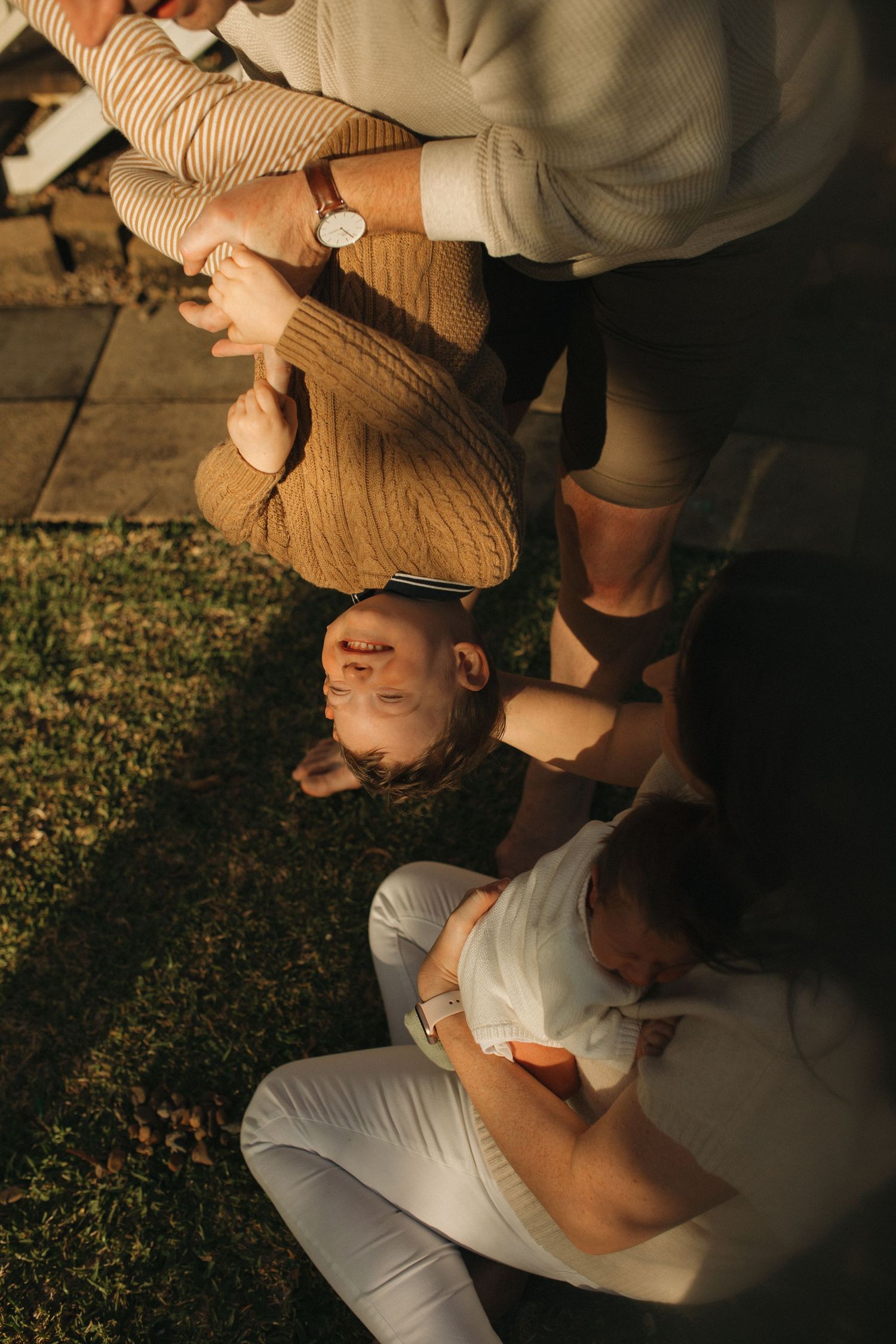 Family sitting on grass in warm sunlight, mother holding baby while child laughs, captured in golden hour light.