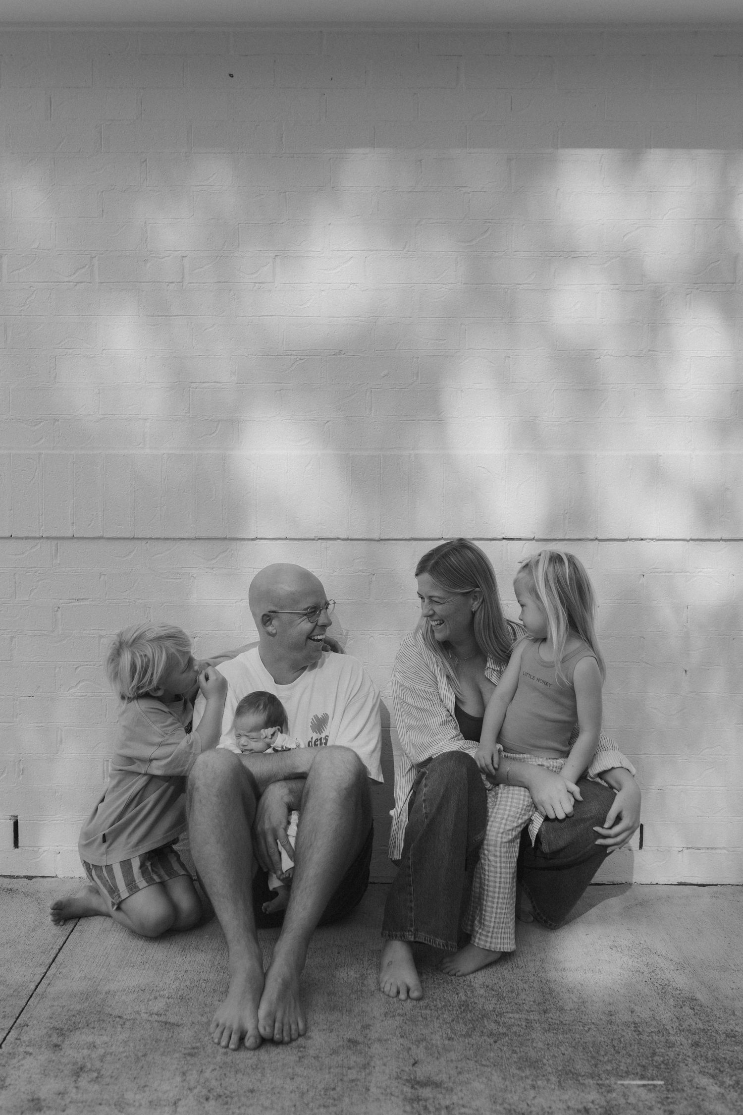 Black and white family photo of parents with three children sitting against a sunlit brick wall outdoors.
