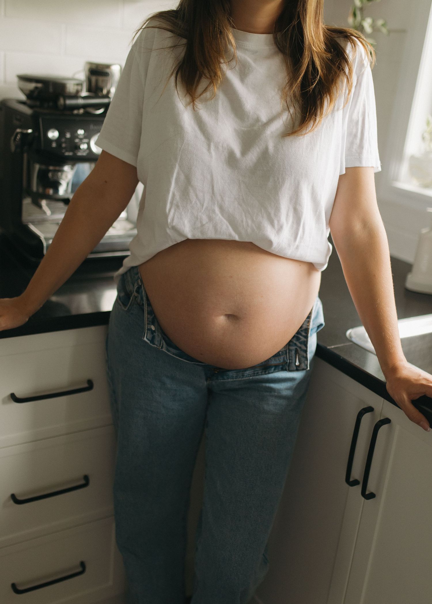 Pregnant woman in white crop top and unbuttoned jeans showing baby bump while standing in modern kitchen. Maternity Photos Brisbane
