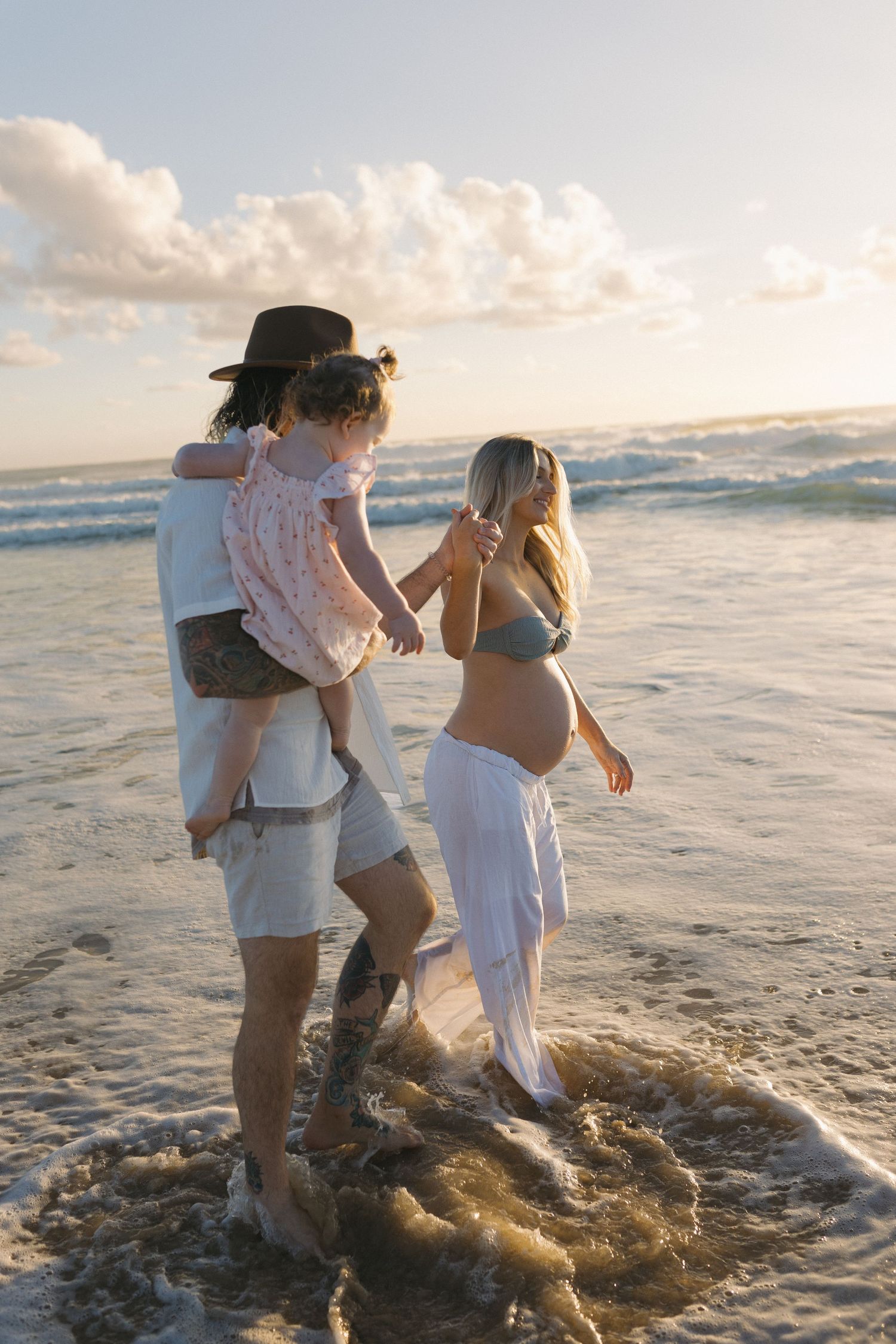 Pregnant woman in bikini top and white pants walks on beach with partner carrying toddler at sunrise.