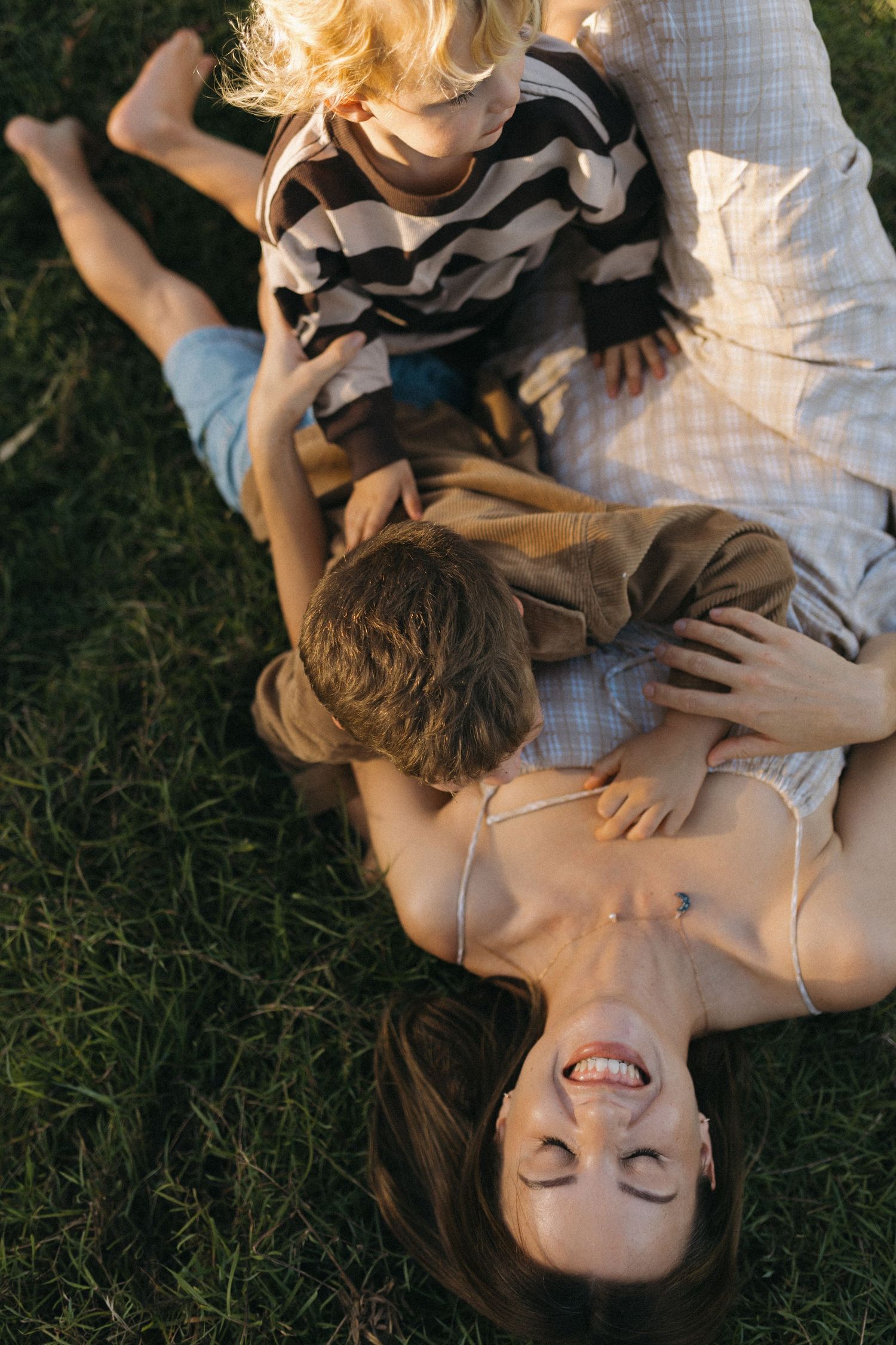 A joyful mother lies on green grass laughing as her two young children playfully cuddle with her outdoors at their property in mount cotton.