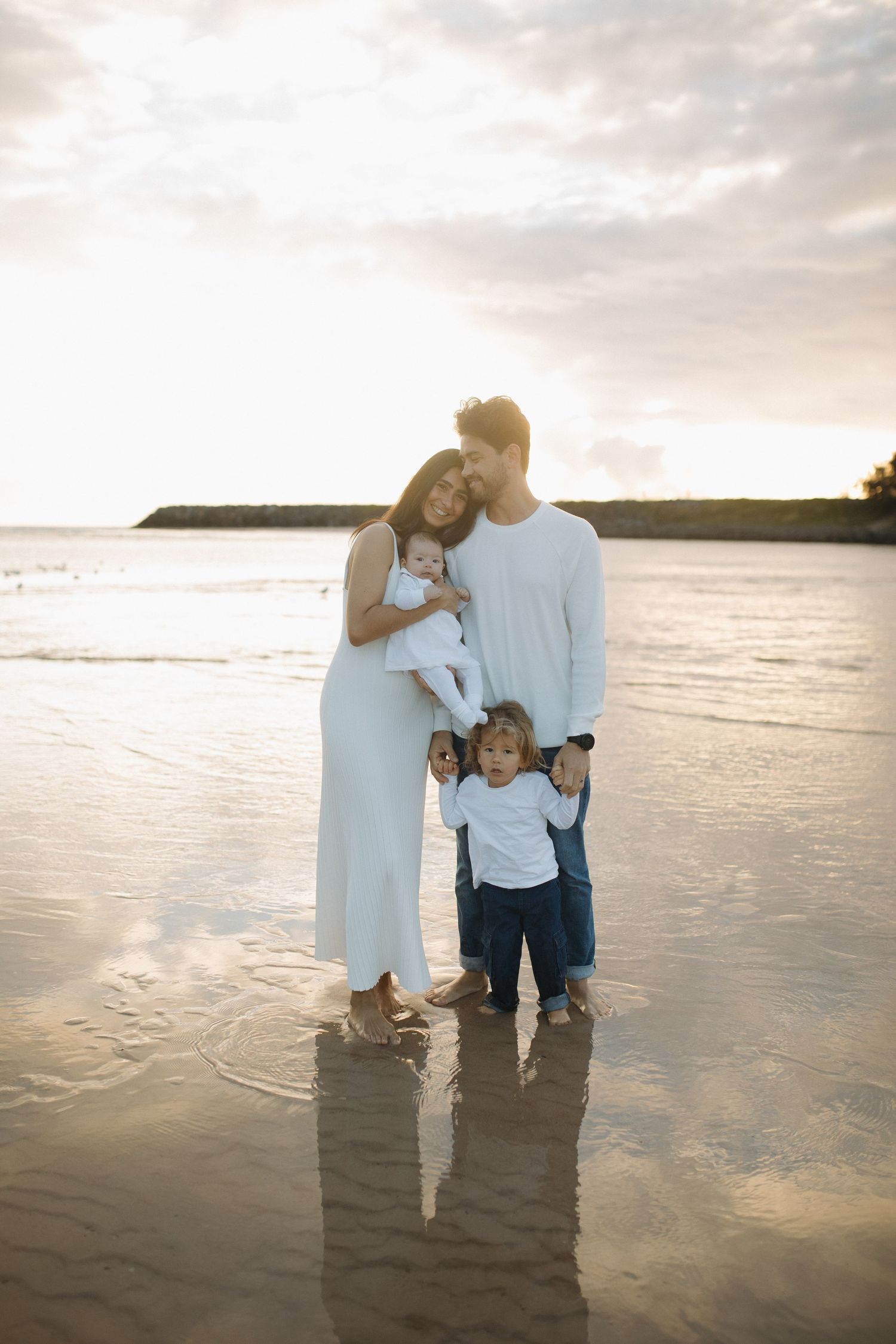 Family of four posing on a beach at sunrise, parents holding a baby while a toddler stands in shallow water.