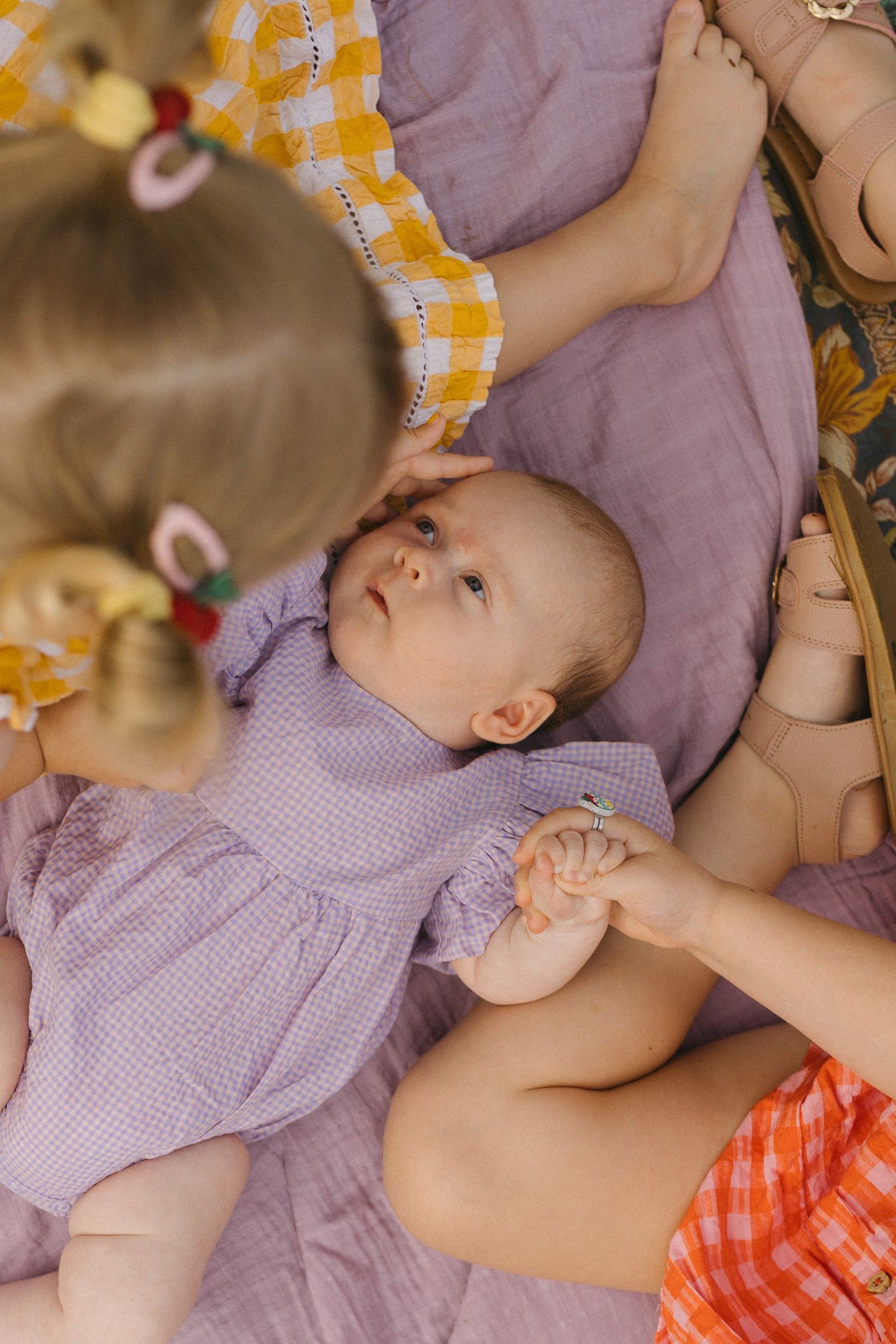 Baby in a purple dress lies on a lilac blanket while a toddler in yellow gingham gently touches her head. In Roma Street Parklands Brisbane