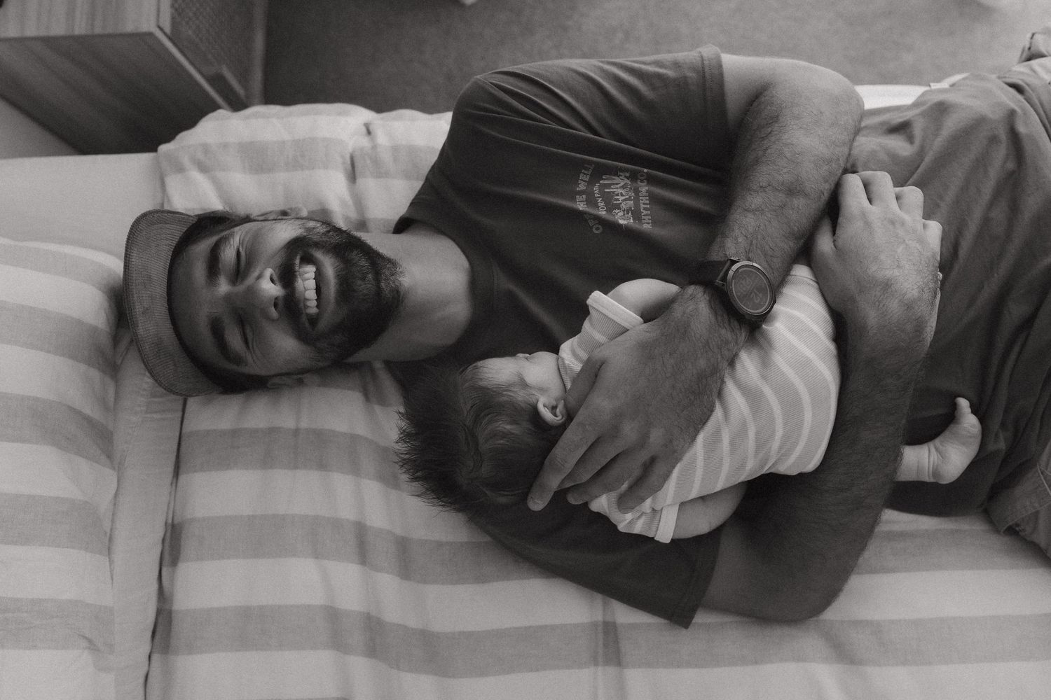 Black and white photo of a bearded man lying on a striped bed, smiling while cradling a newborn baby.