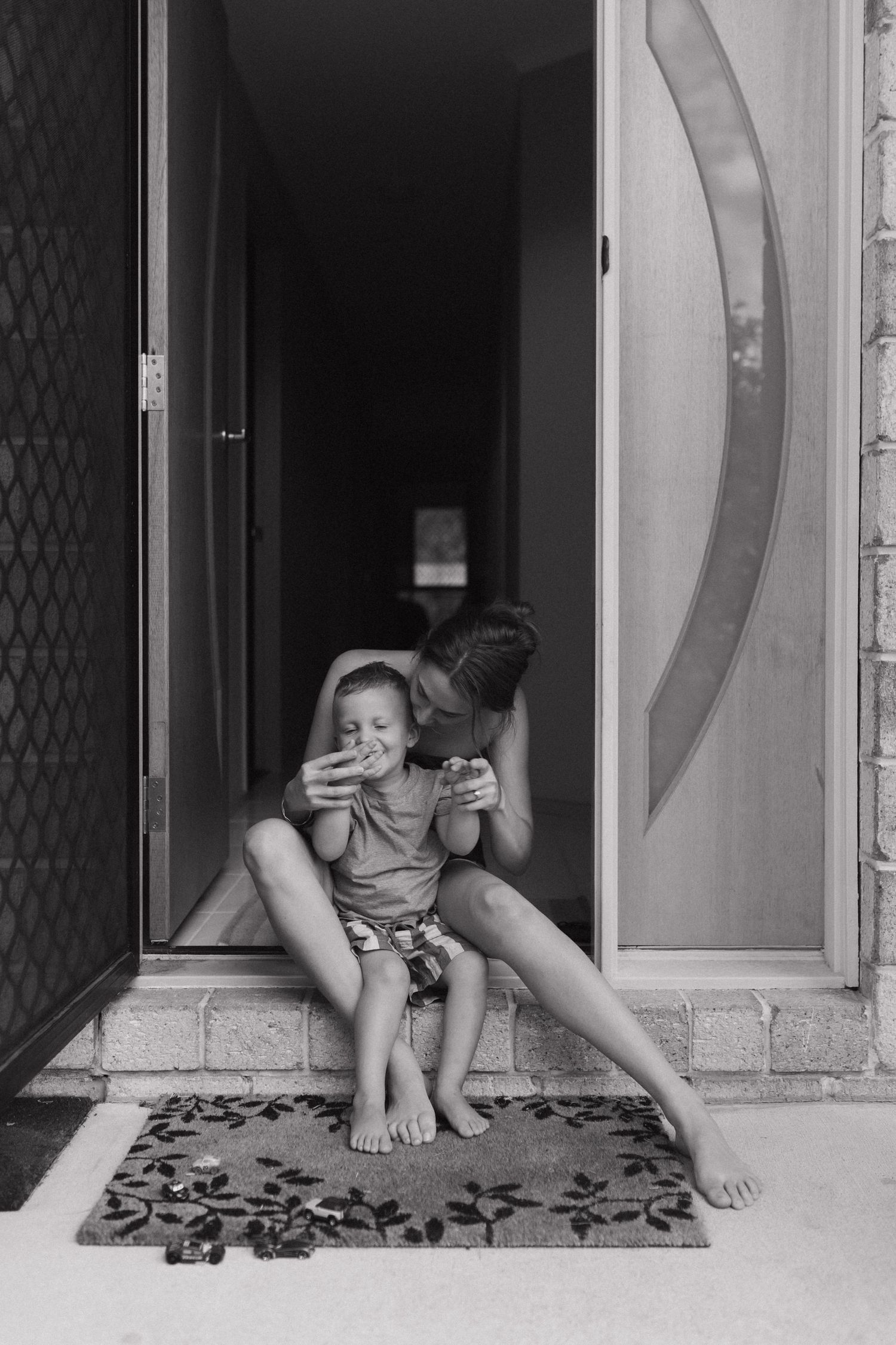 Black and white photo of a mother and young child sitting on a doorstep, both having cuddle. In North Brisbane in home photography session
