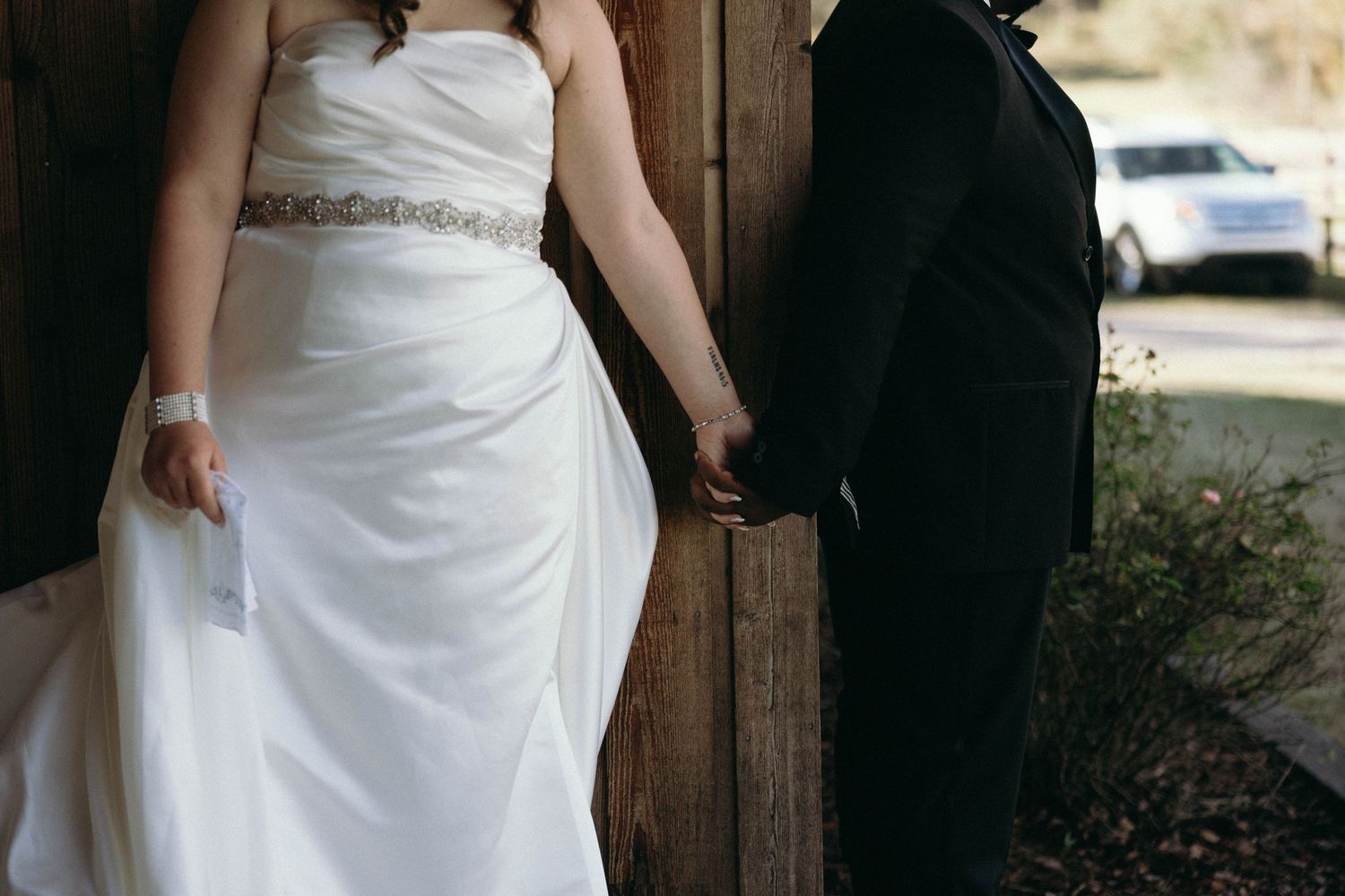 A close-up view of a bride in white dress holding hands with groom in black suit near wooden post.