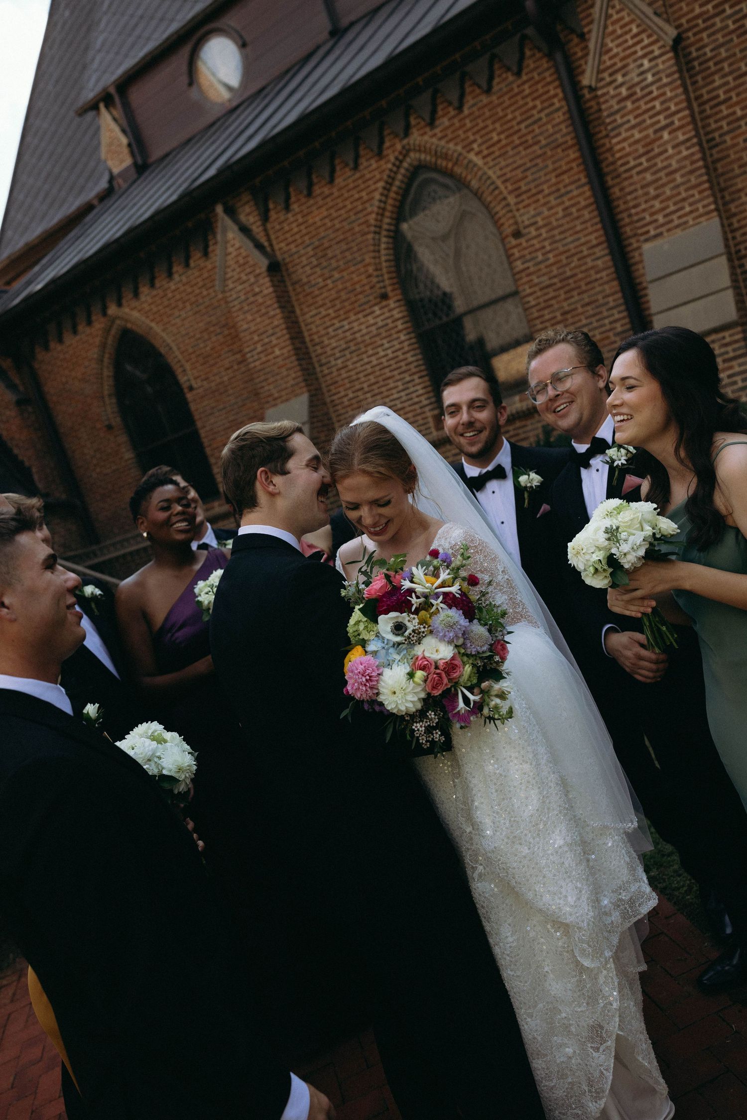 A wedding party shares a joyful moment together outside a brick church with gothic architecture.