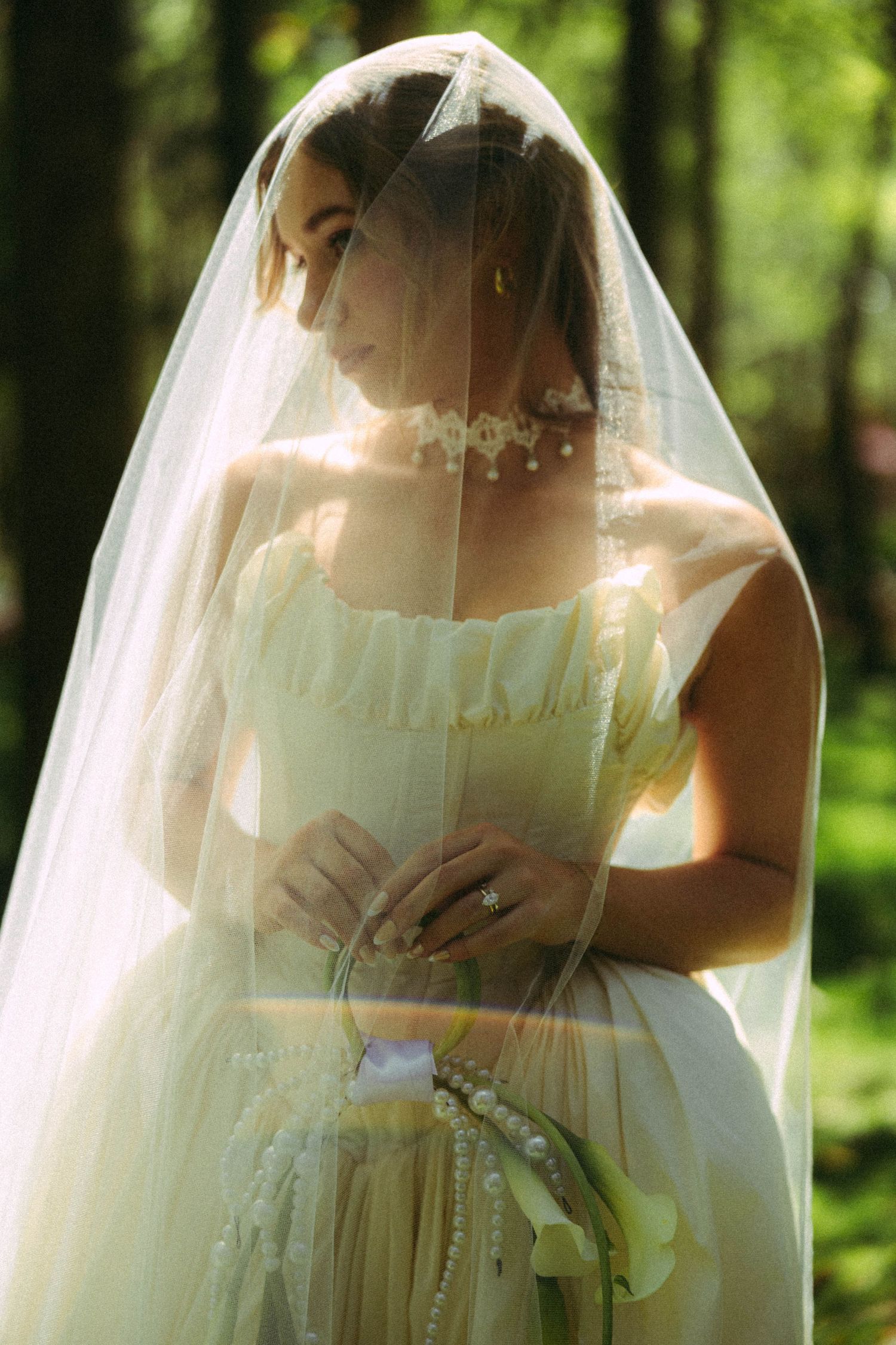 Bride in ivory gown and veil holding pearl bouquet in sunlit forest setting.