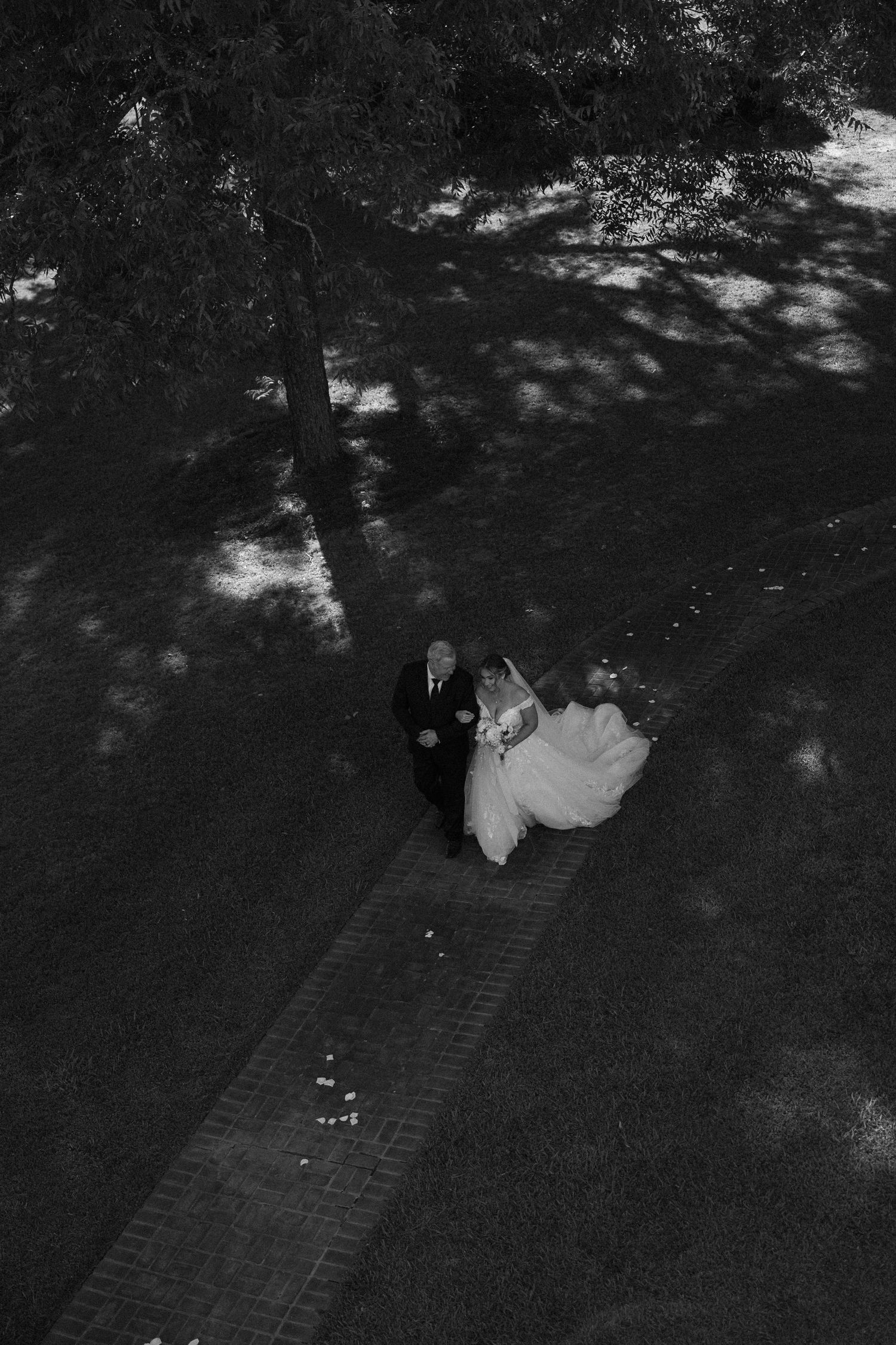 Aerial black and white view of a couple walking down a brick path surrounded by trees and dappled shadows.