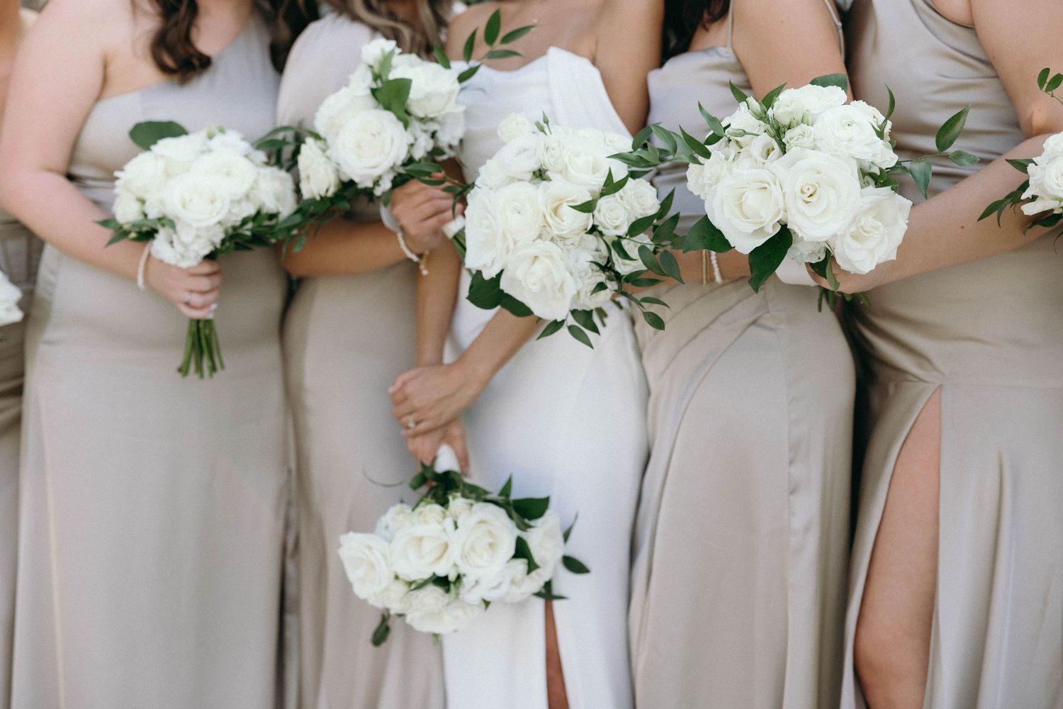 Bridesmaids in beige dresses hold elegant white peony bouquets during a wedding ceremony.