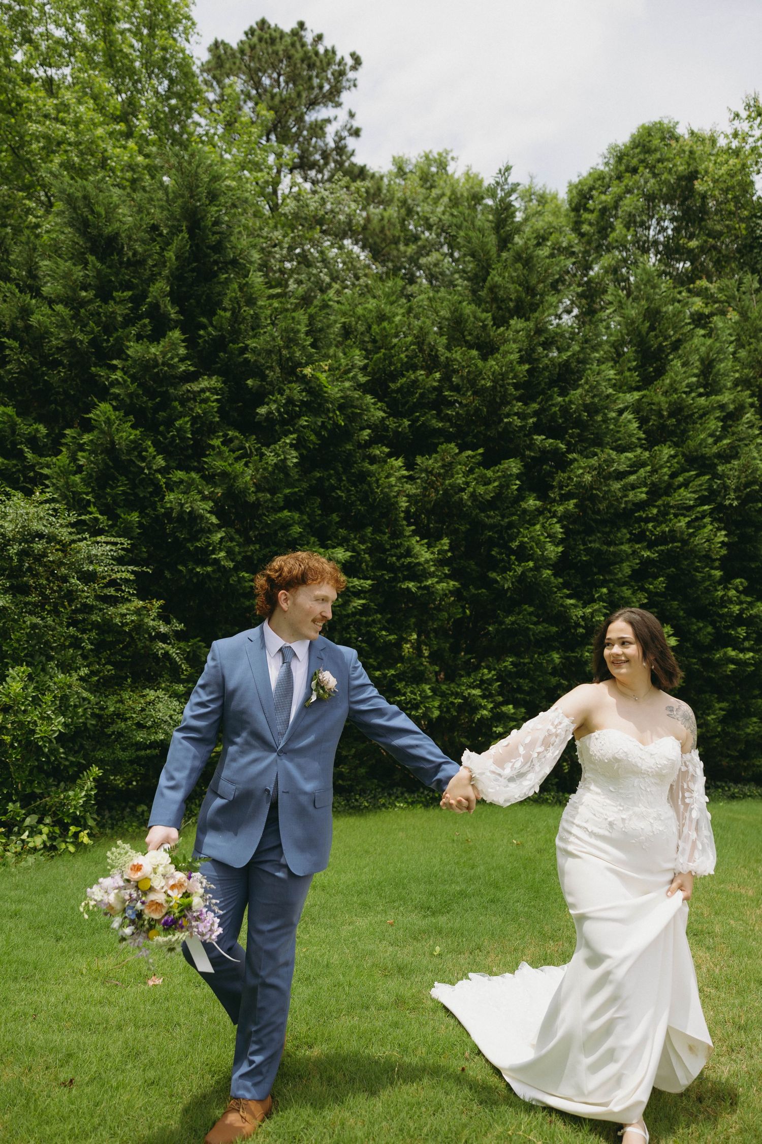 A bride and groom hold hands walking across green grass with lush trees behind them on their wedding day.