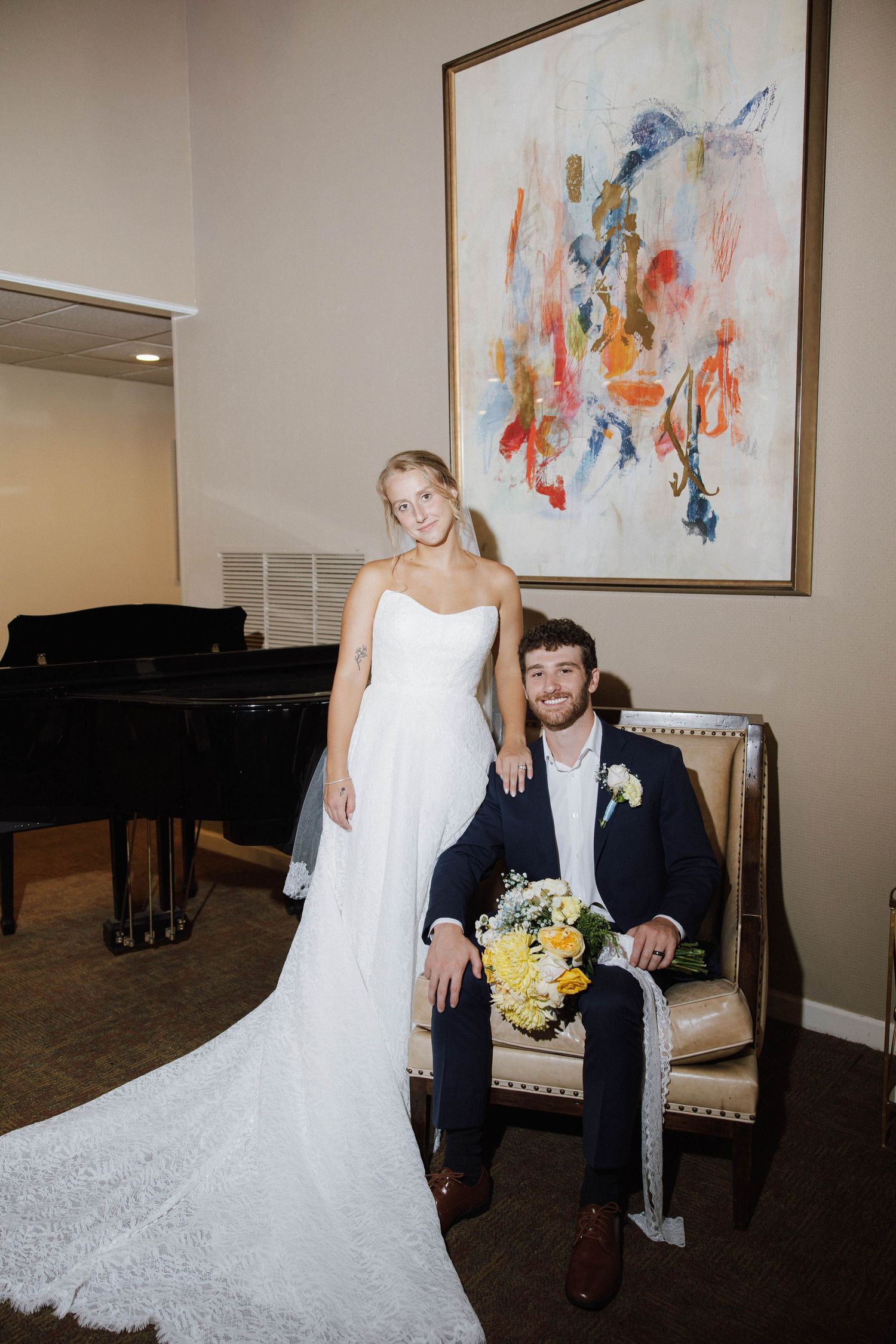 Bride in white gown stands beside groom in navy suit seated in chair, holding yellow bouquet before colorful abstract art.