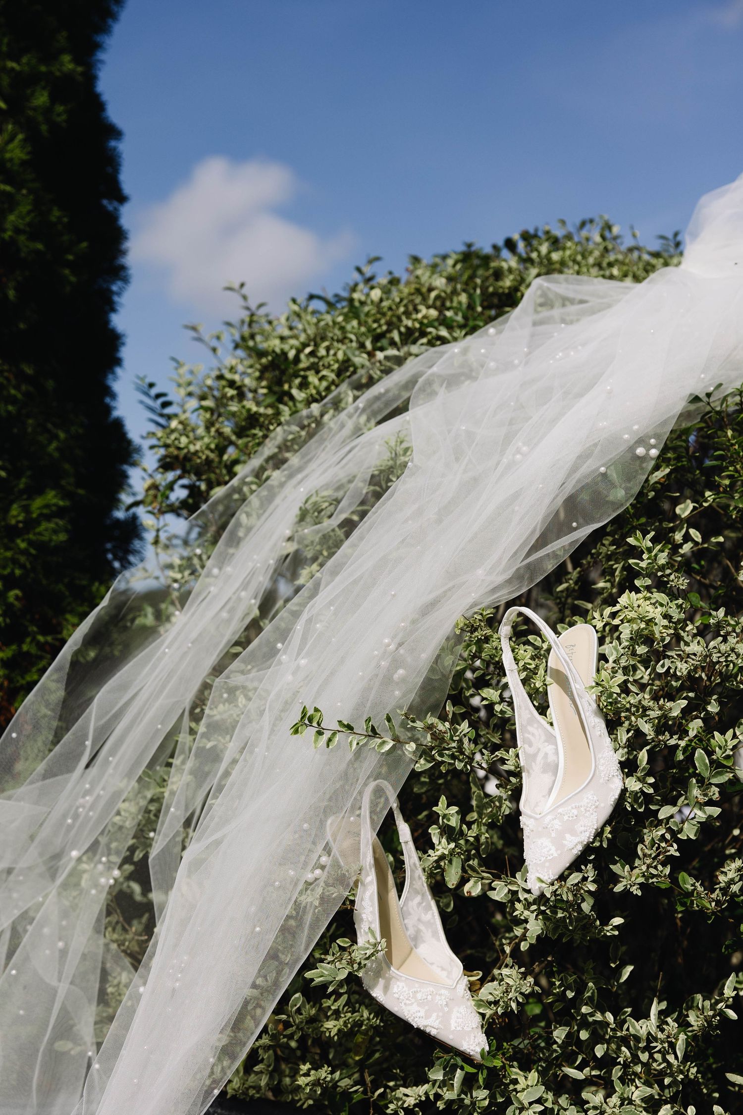 White bridal veil and elegant slingback heels displayed against green garden foliage on a sunny day.