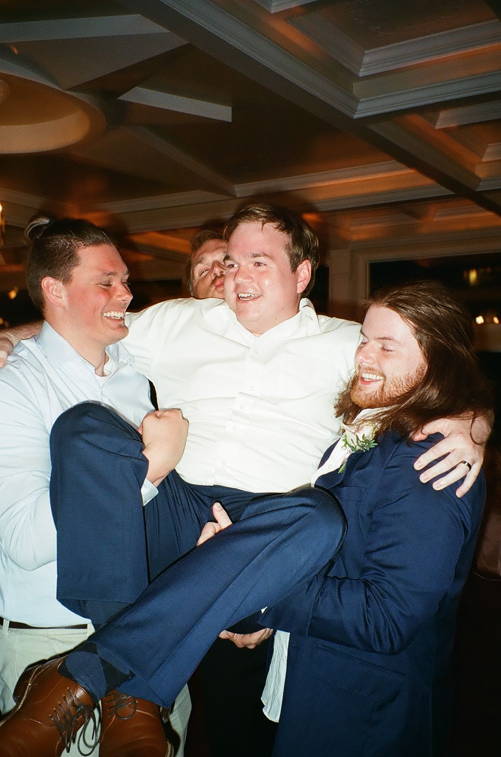 Three friends share a joyful moment at a party as two carry their companion in navy suits and white shirts.