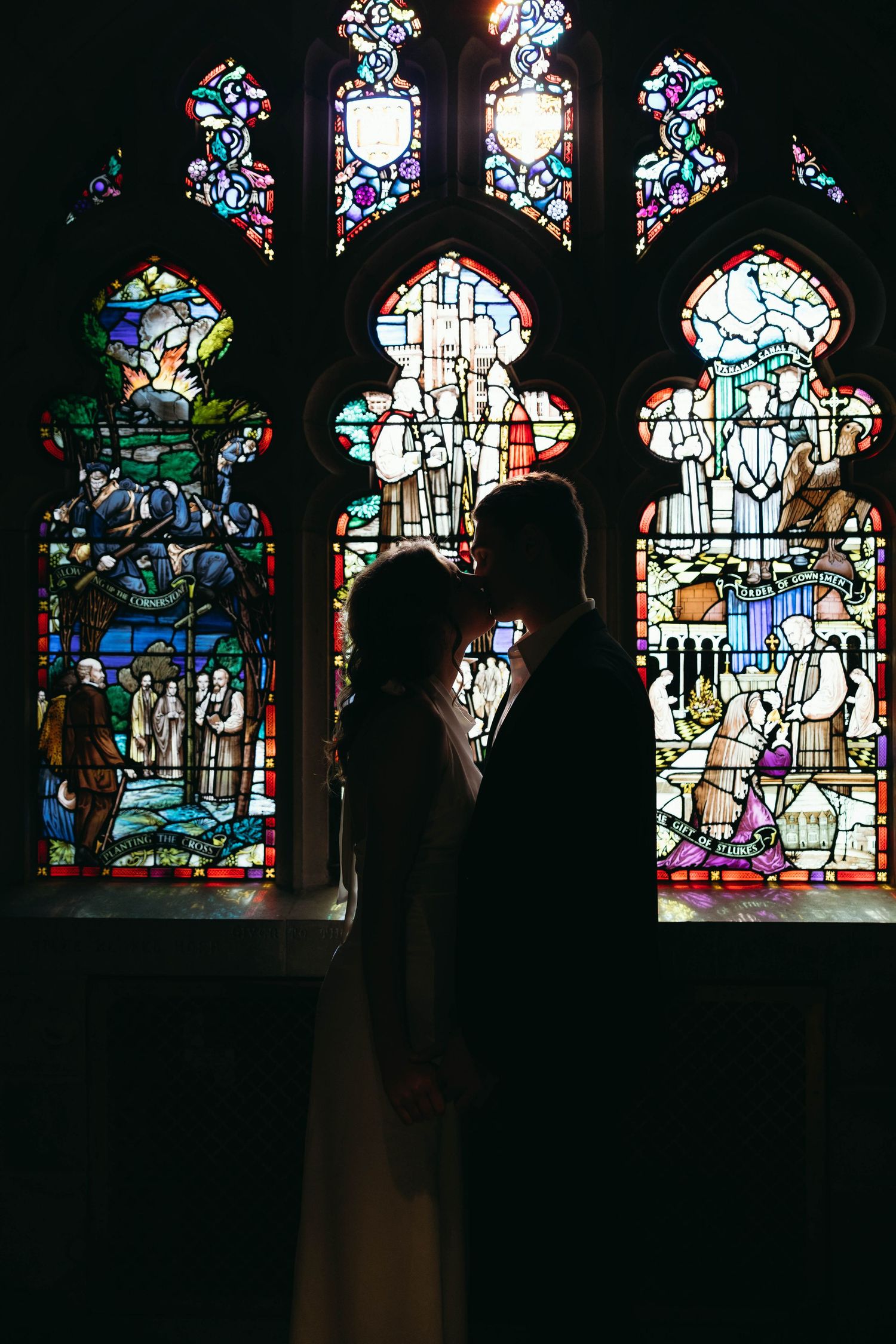 Silhouette of bride and groom sharing a kiss in front of stunning colorful stained glass church windows.
